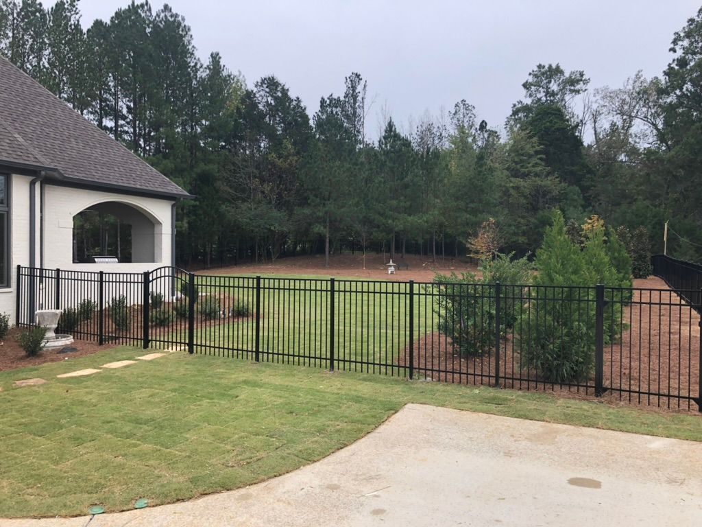 Black metal fence surrounding a yard with a house, grass, and trees.