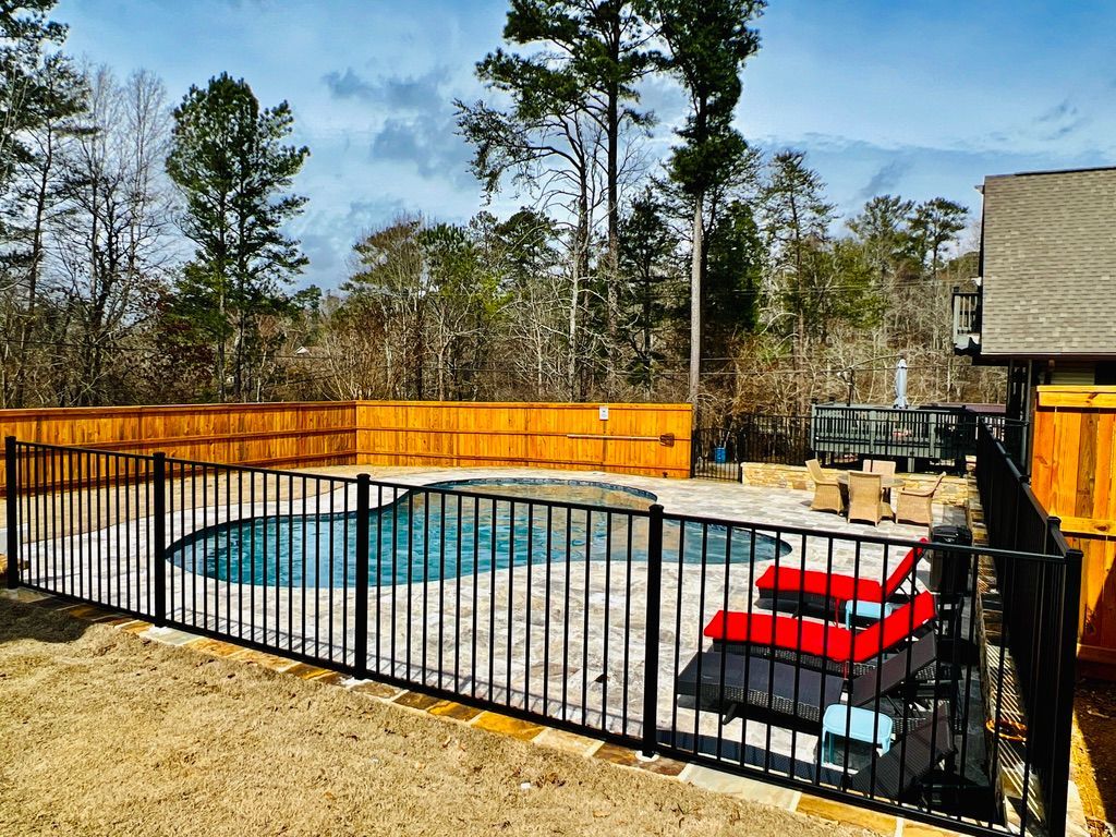 Black fenced pool with lounge chairs and surrounding stone patio, with wooden fence and trees in background.