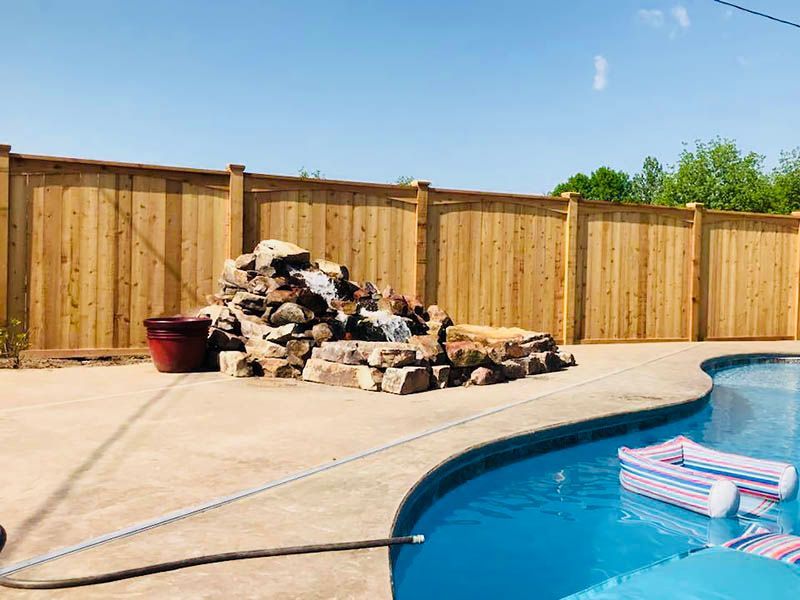 Backyard pool with wooden fence and a rock waterfall feature.