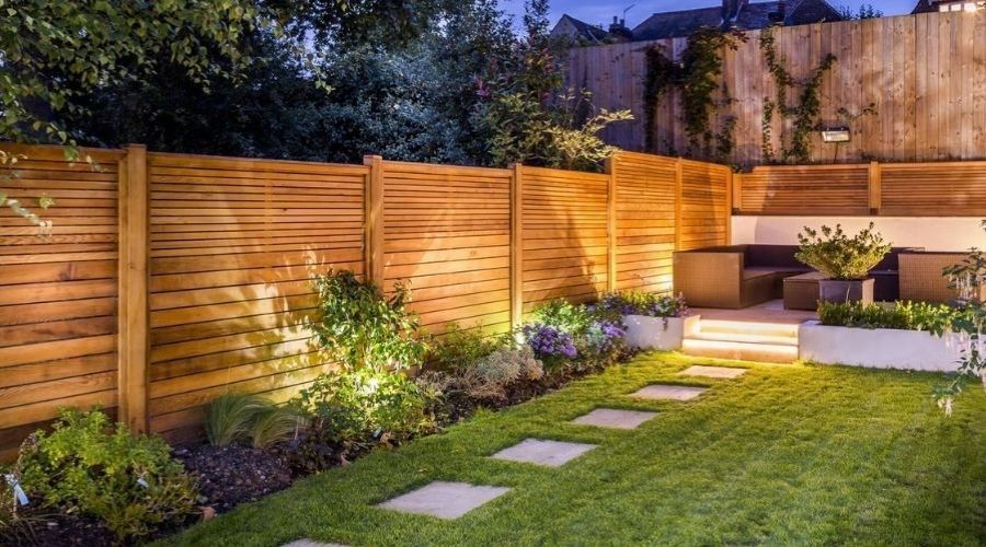 Stepped horizontal wooden privacy fence enclosing a sloped, terraced garden with evening lighting