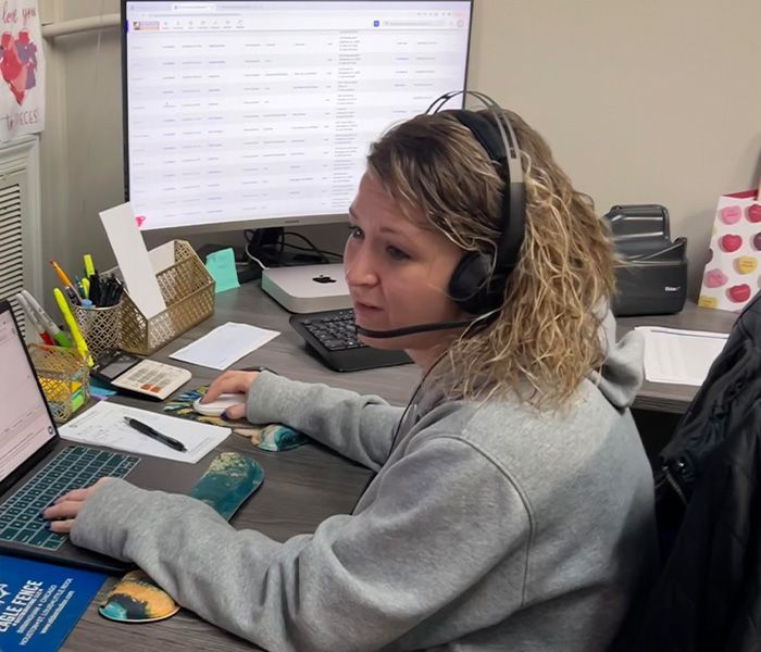 Woman wearing headset, working at desk with laptop, monitor.