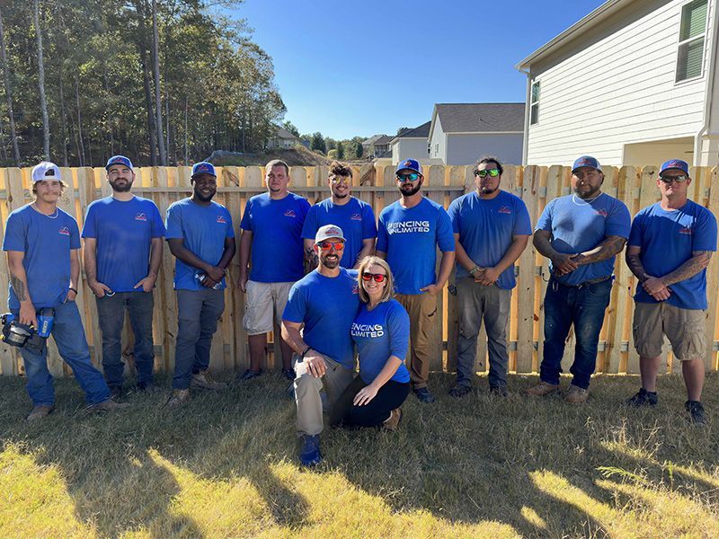 A group of people wearing blue shirts posing in front of a wooden fence on a sunny day.