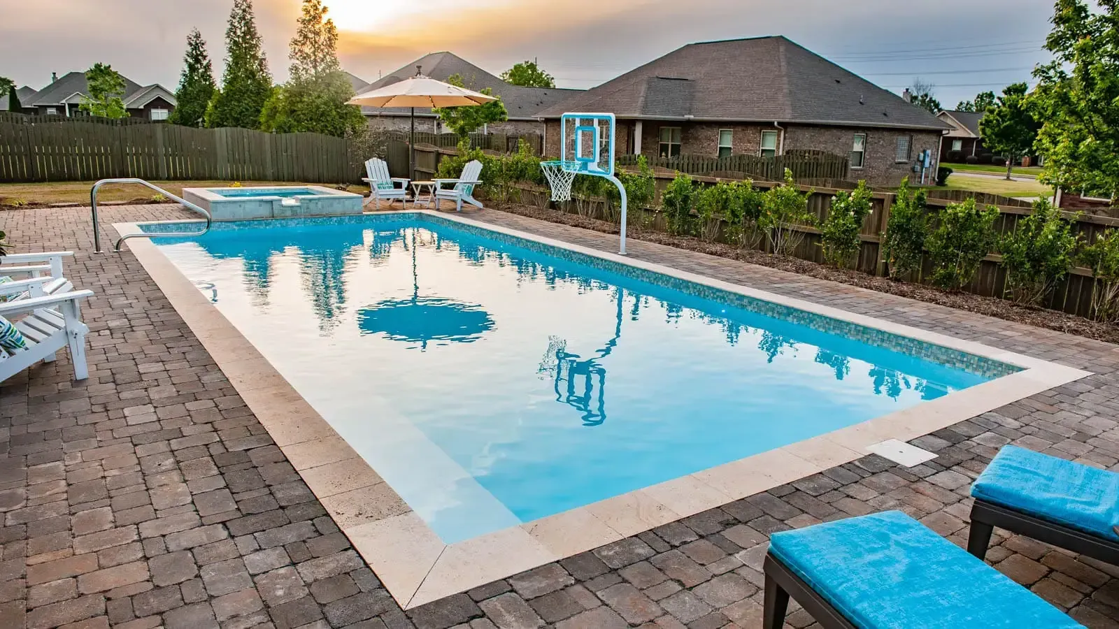 Backyard swimming pool with turquoise water, stone patio, lounge chairs, and houses at sunset