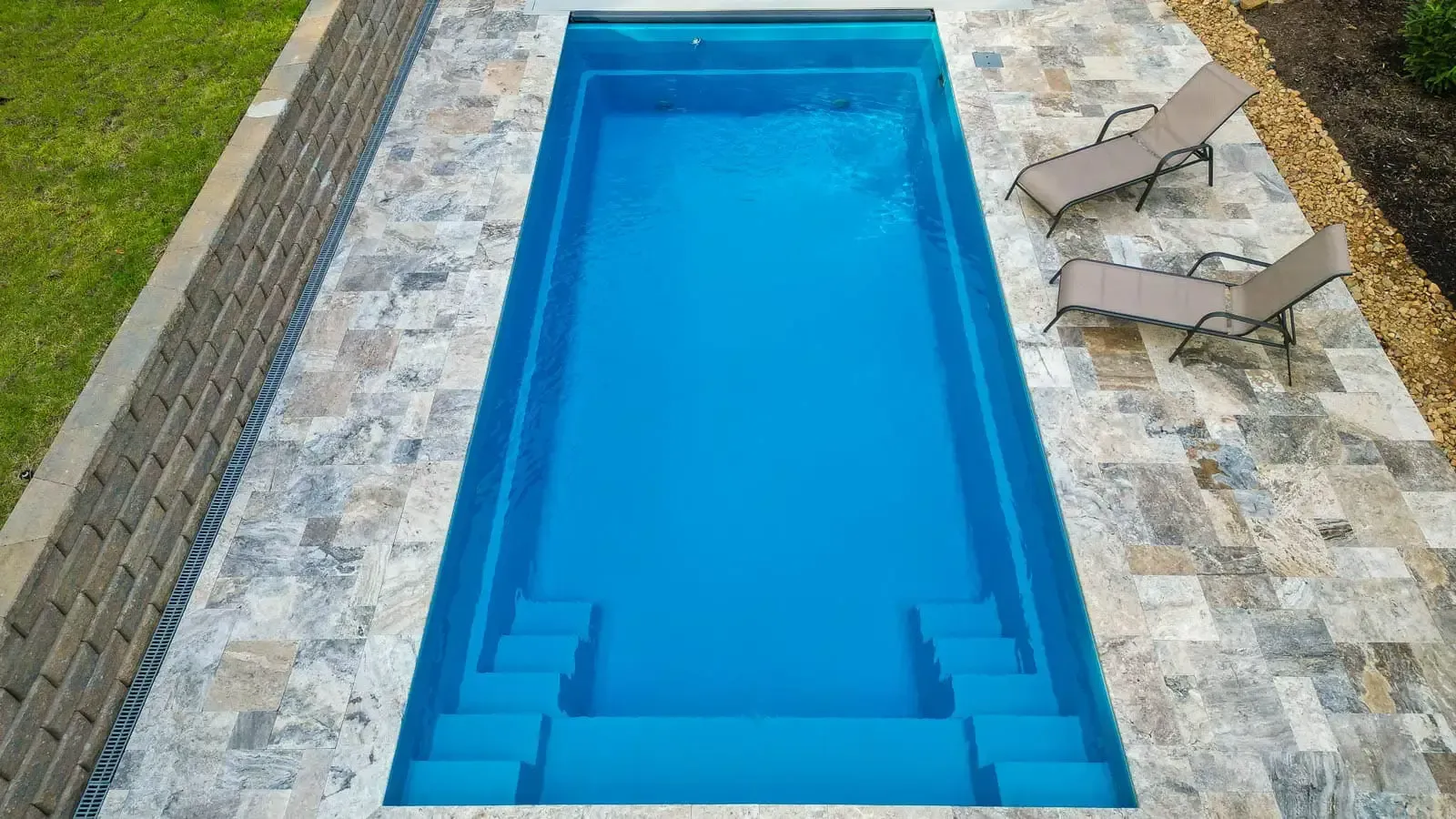 Rectangular blue swimming pool with stone deck and lounge chairs at the top right