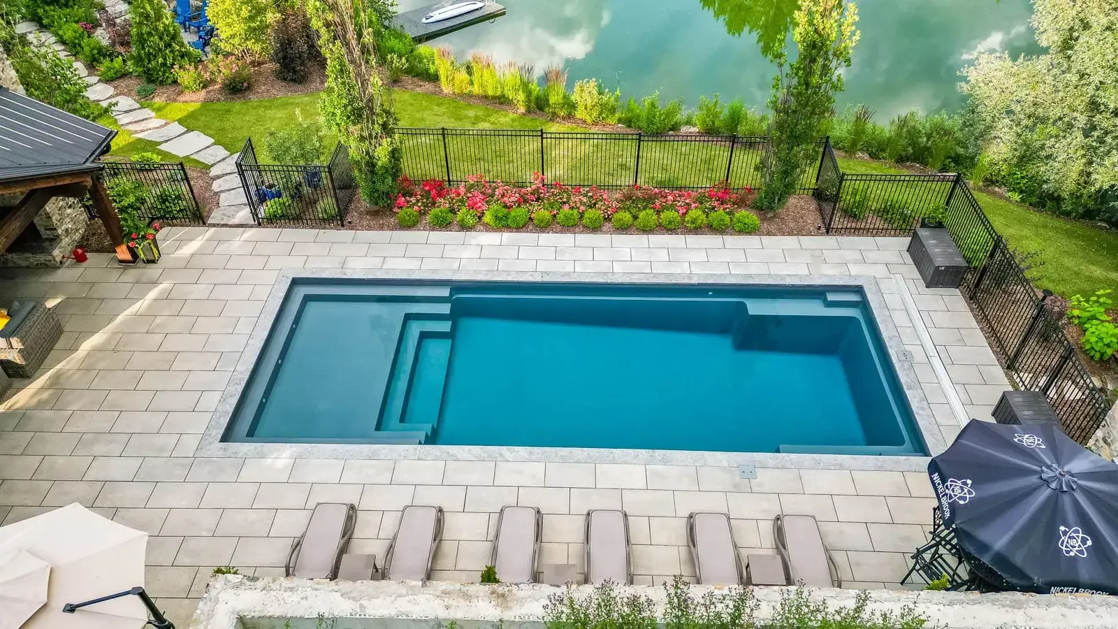 Rectangular backyard pool with stone patio, green landscaping, and a lakeside view