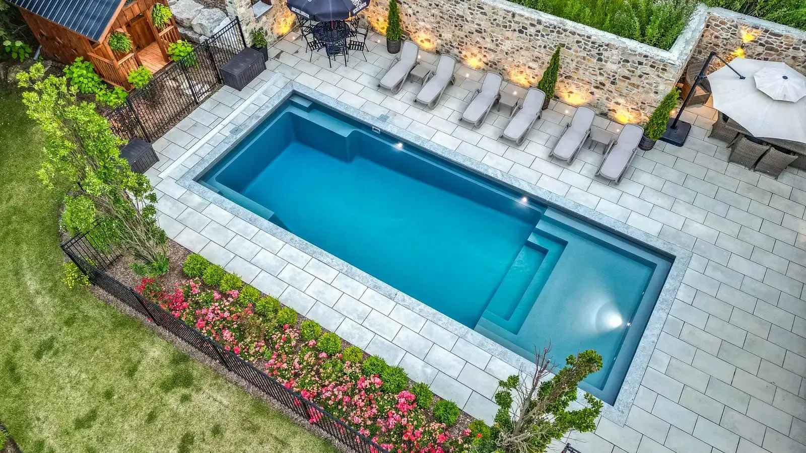 Overhead view of a small rectangular backyard pool with lounge chairs and stone patio surrounded by flowers and greenery