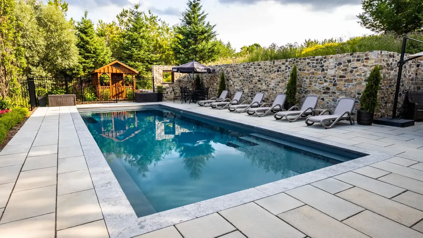 Backyard pool with lounge chairs, stone wall, and a gazebo surrounded by trees