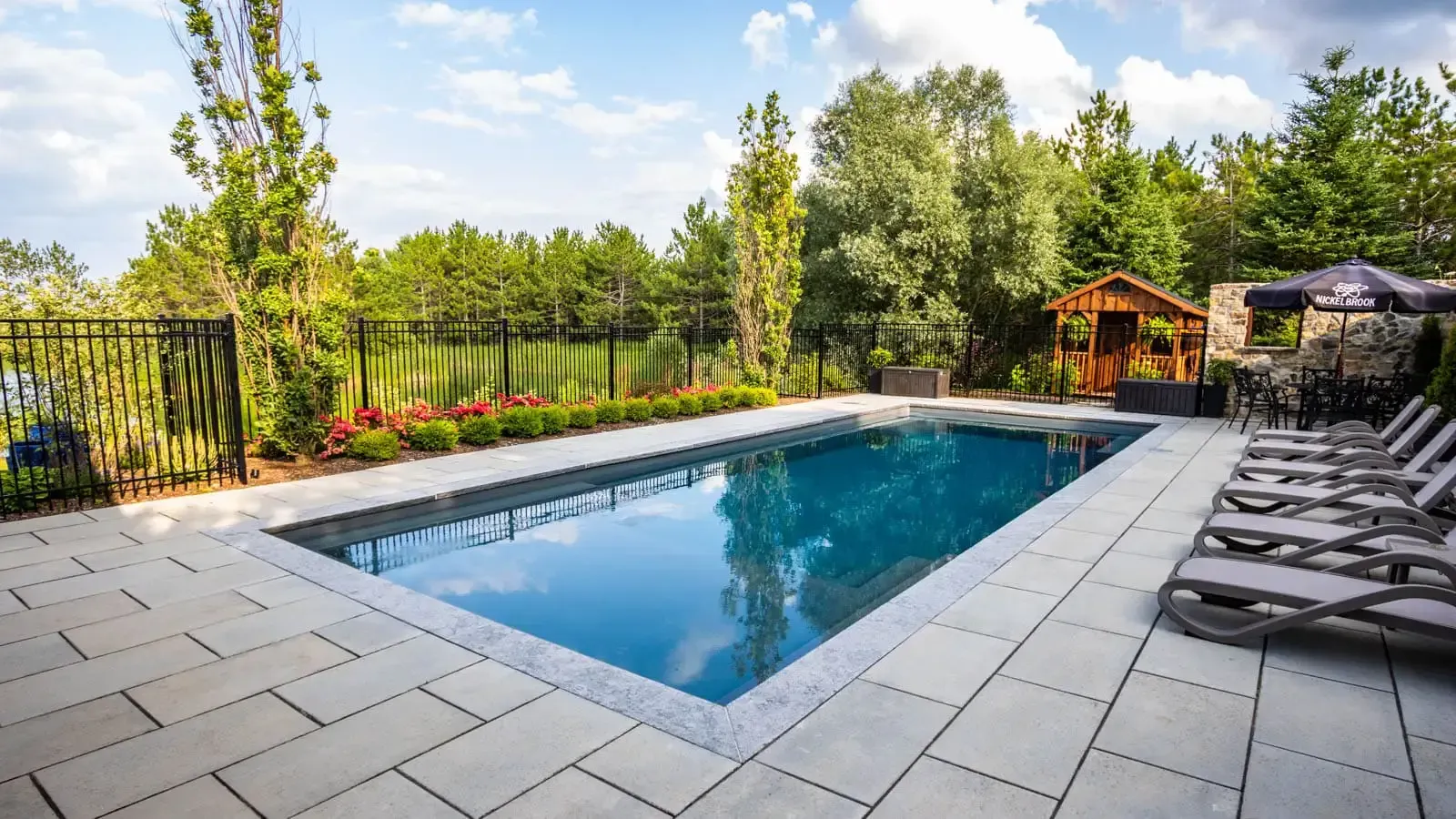 Backyard patio with a blue swimming pool, lounge chairs, and trees in the background