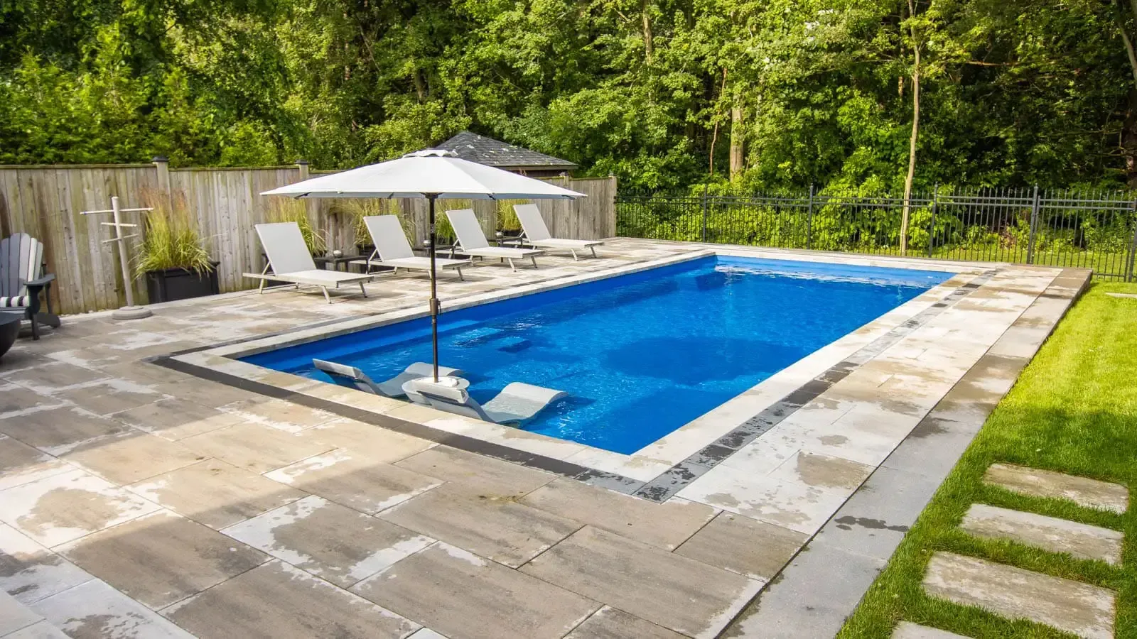 Backyard swimming pool with blue water, patio, umbrella, and lounge chairs surrounded by trees