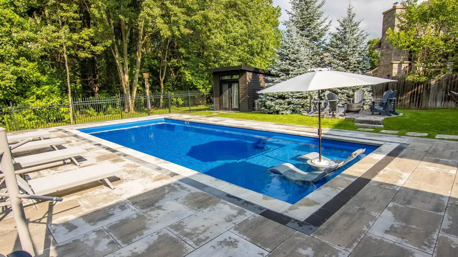 Backyard pool with lounge chairs, white umbrella, and trees surrounding a blue tiled water feature