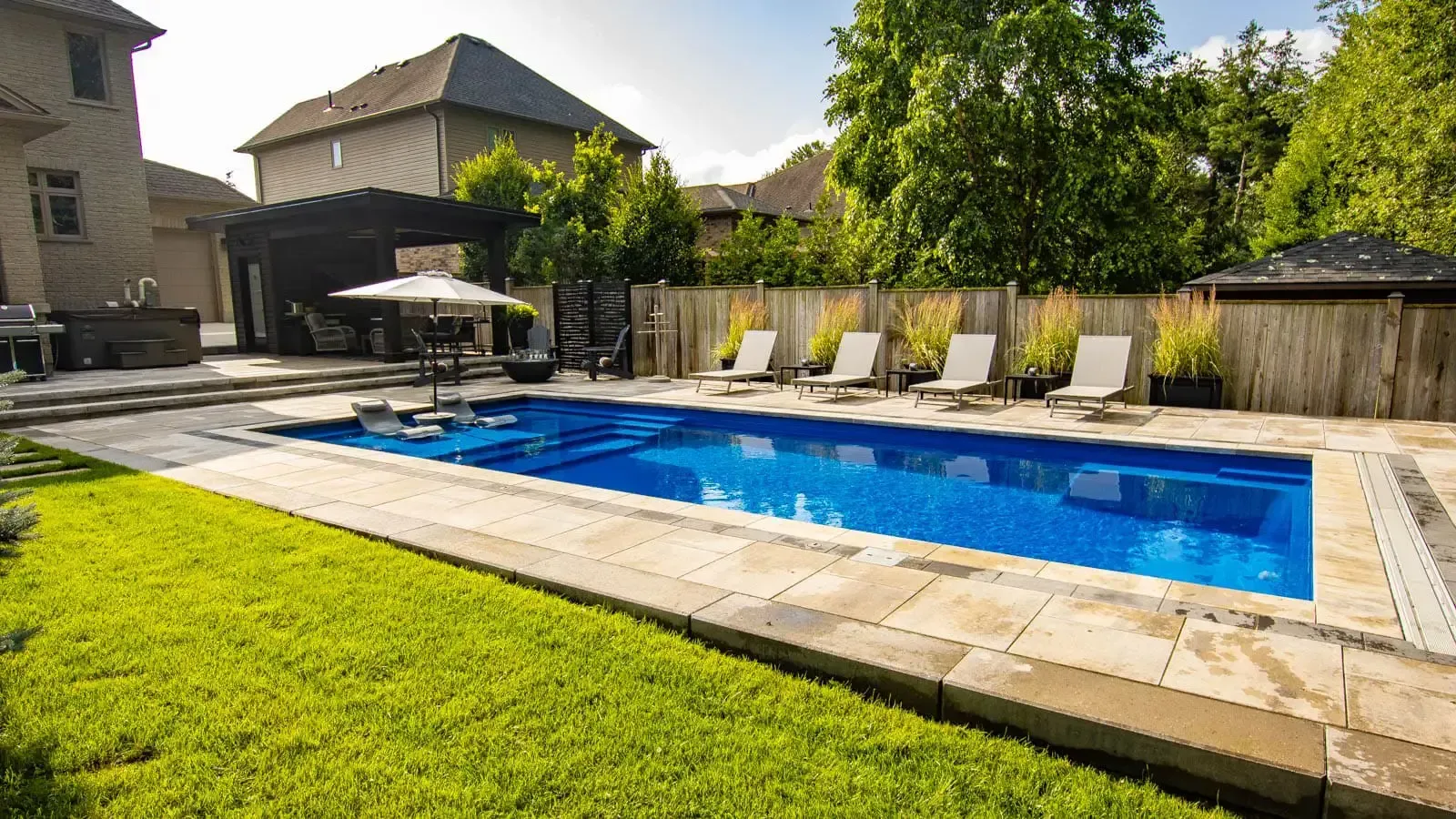 Backyard swimming pool with lounge chairs, patio umbrellas, and green lawn in sunlight