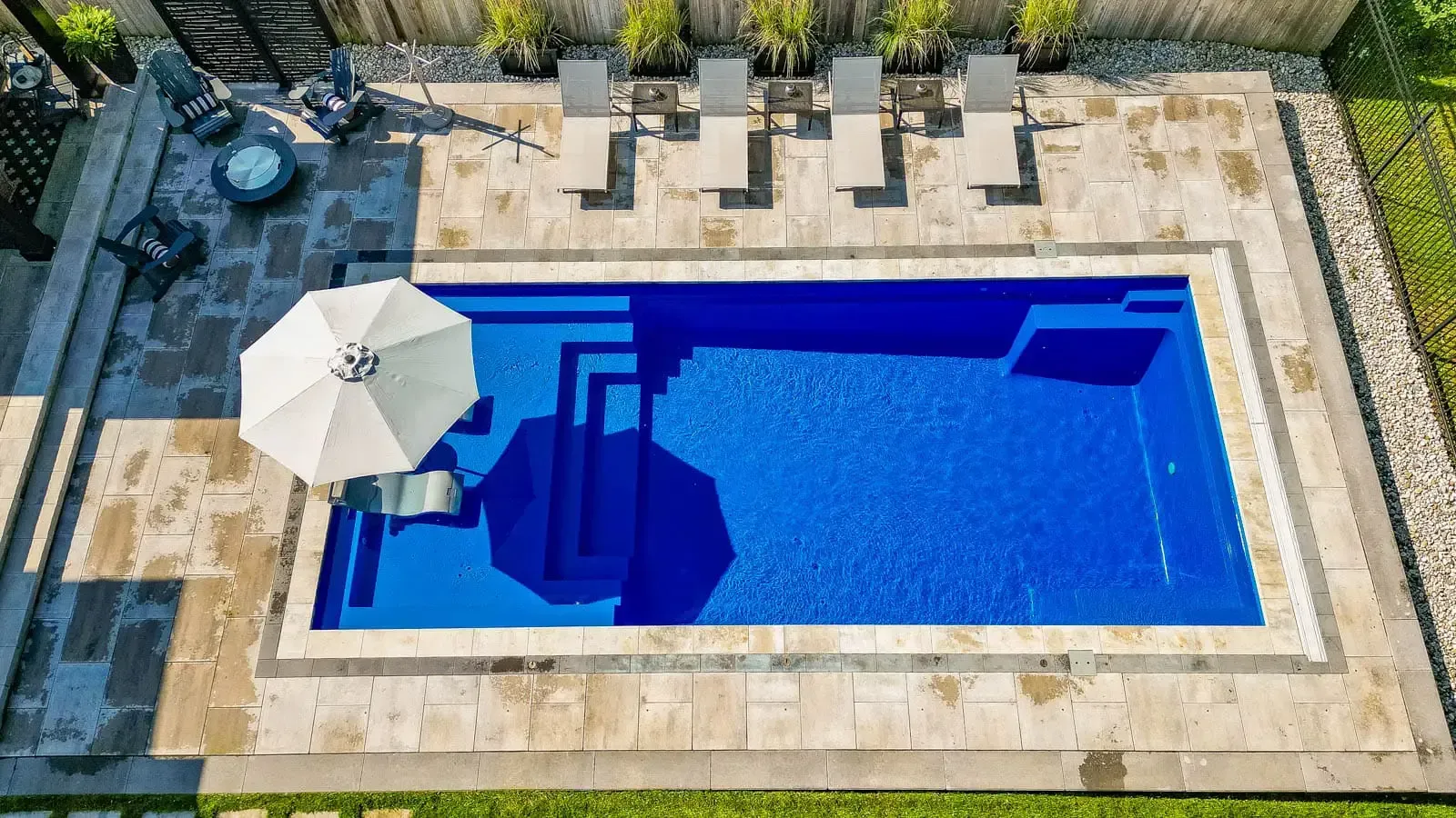 Overhead view of a rectangular blue swimming pool with beige deck chairs and a white umbrella