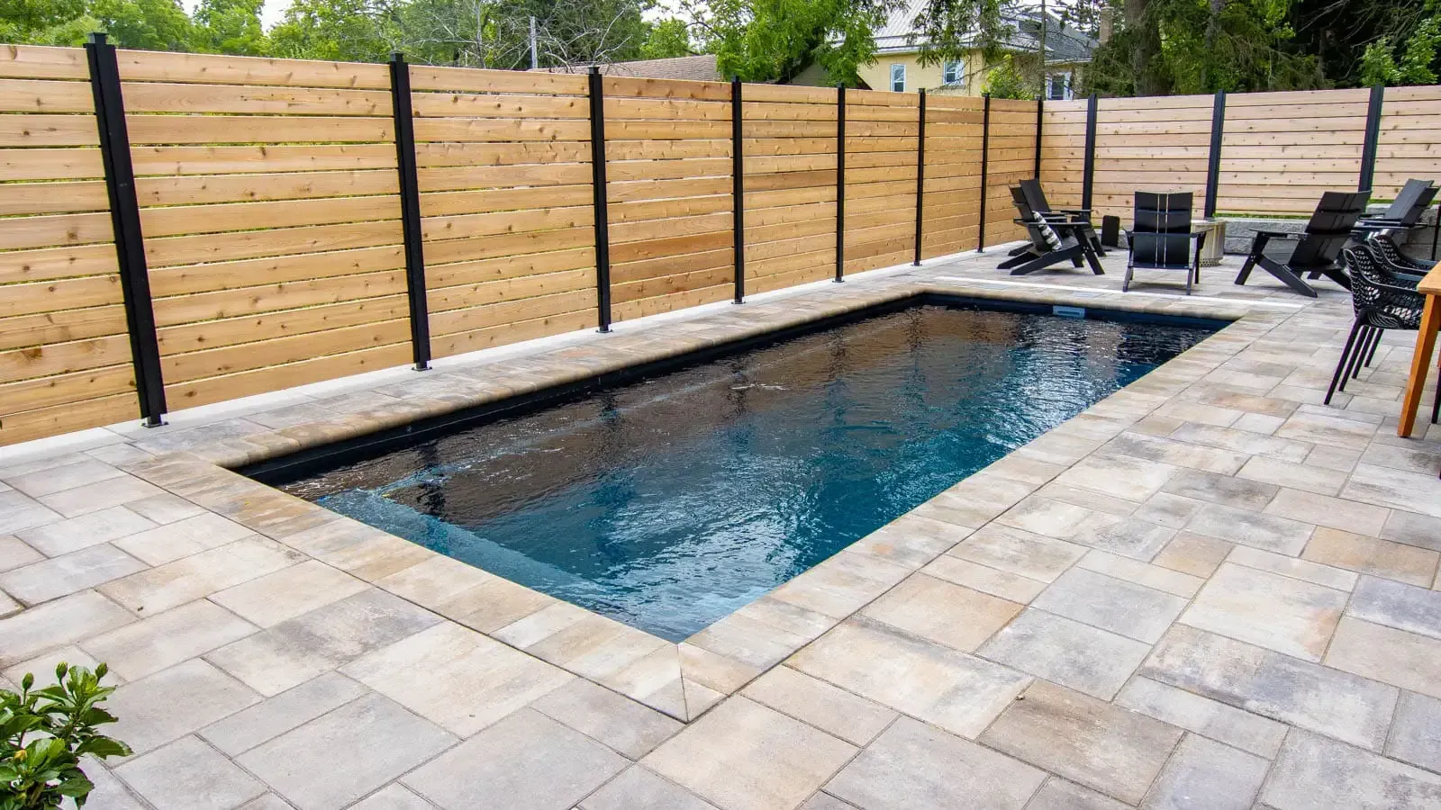 Rectangular backyard pool with stone patio, wooden fence, and lounge chairs by the water