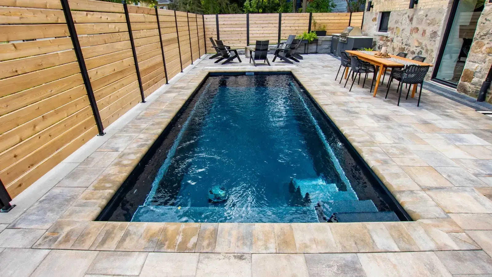 Rectangular backyard pool with dark blue water, stone deck, lounge chairs, and wooden privacy fencing