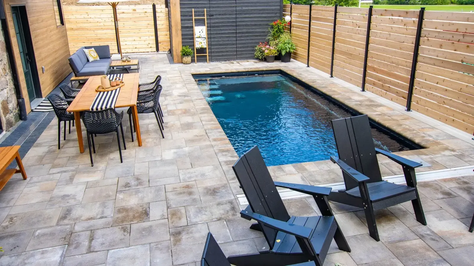 Modern backyard with a narrow swimming pool, stone patio, and black lounge chairs beside a dining table