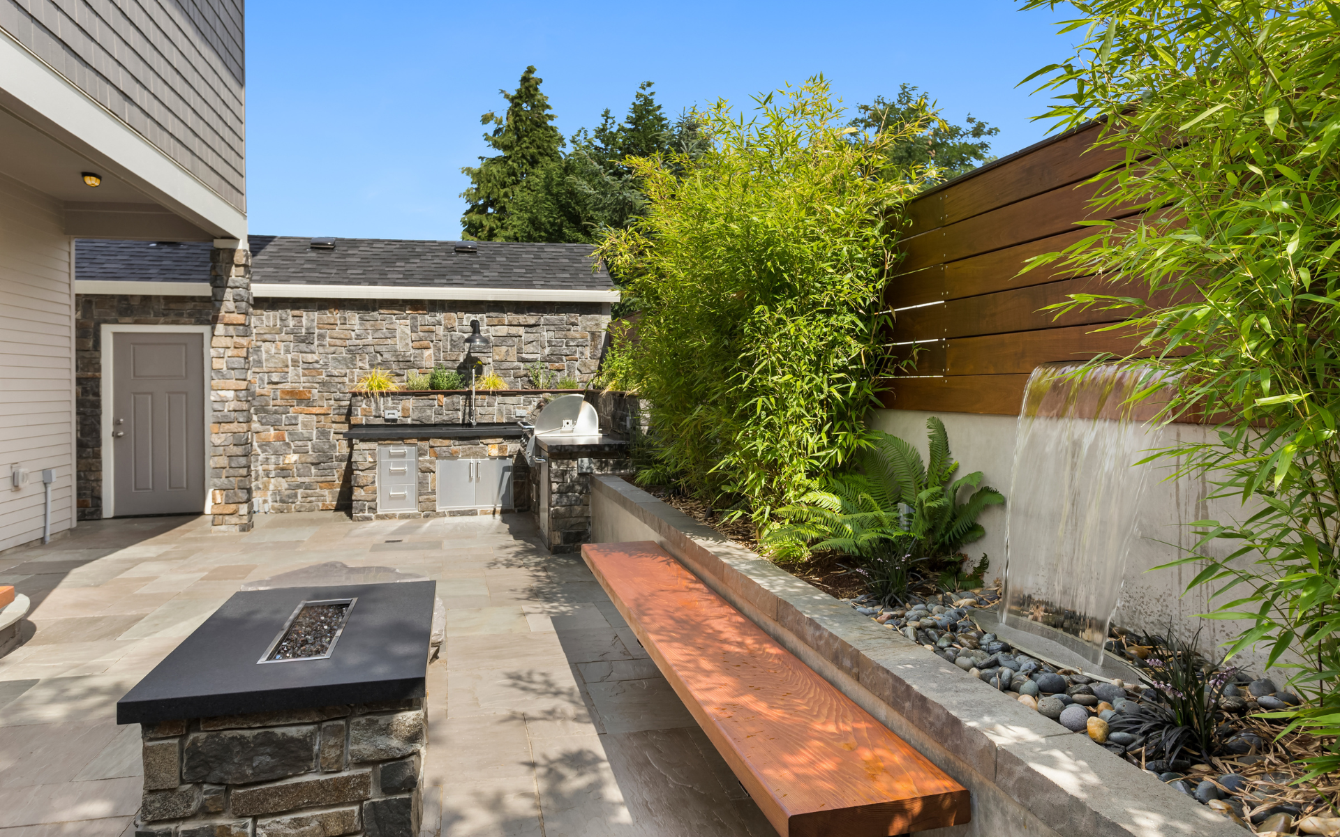 Modern patio with stone wall, built-in fire pit, wooden bench, and greenery along a concrete walkway