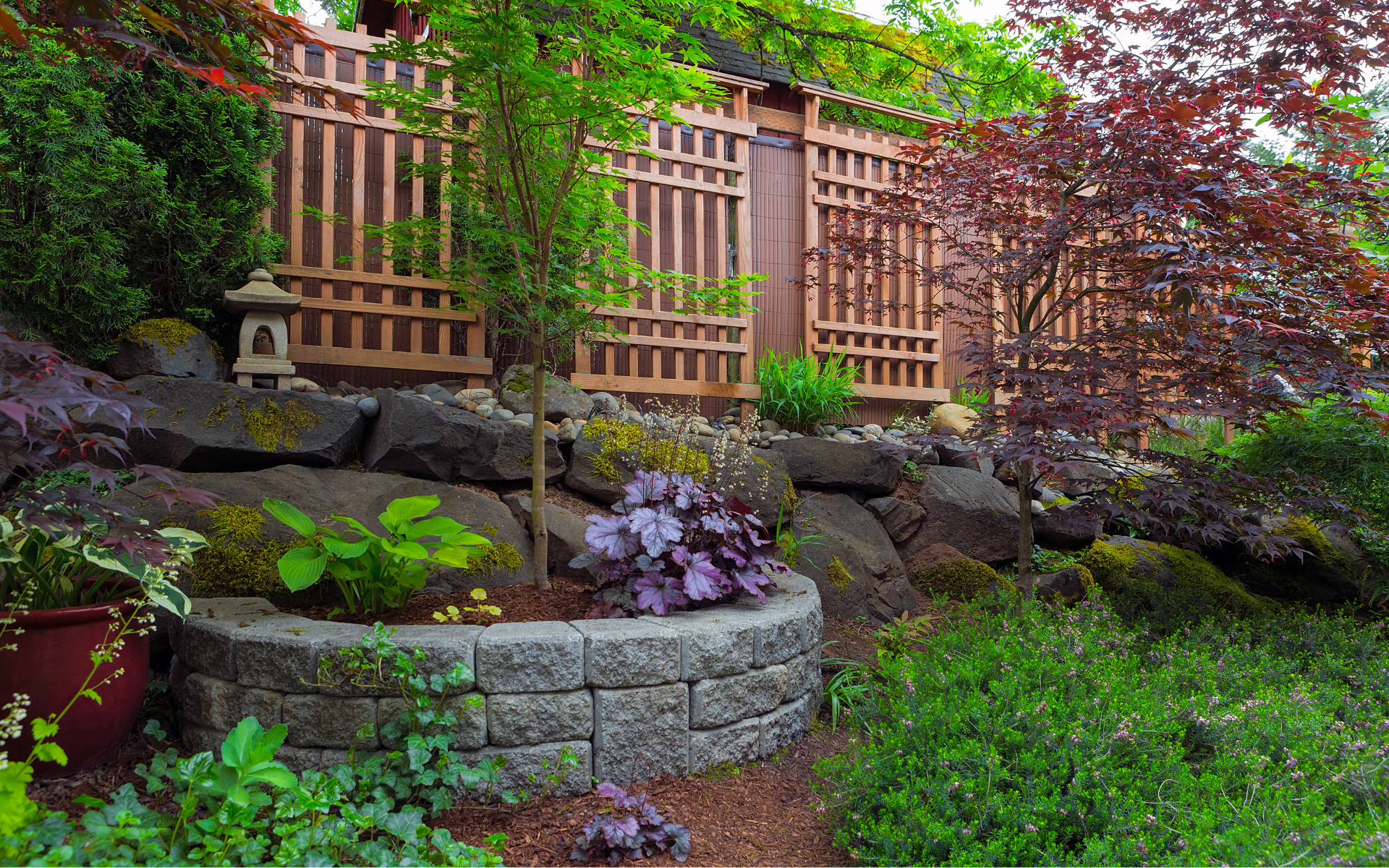Lush garden with stone retaining wall, purple flowers, and wooden fence behind greenery.
