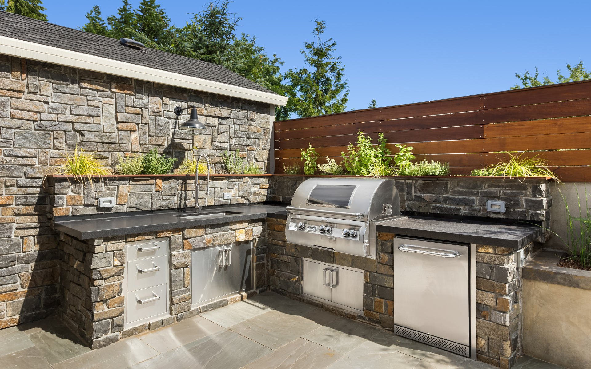 Outdoor stone backyard kitchen with built-in grill, sink, and cabinets under a wooden fence and blue sky