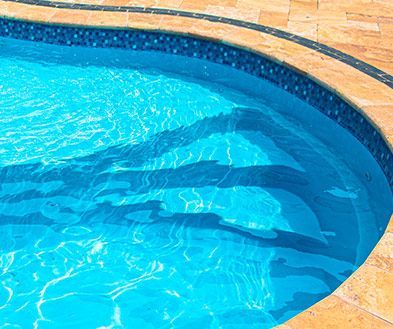Curved blue swimming pool with sunlit water and beige tile deck کنار