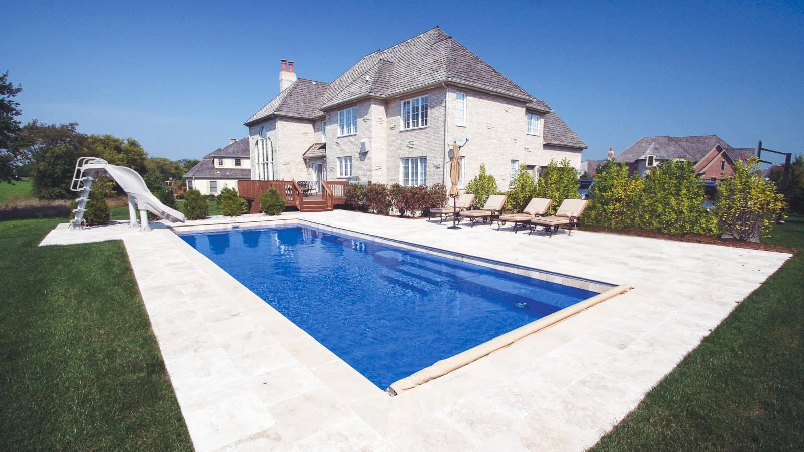 Luxury home with a rectangular blue swimming pool and surrounding white patio in a sunny yard