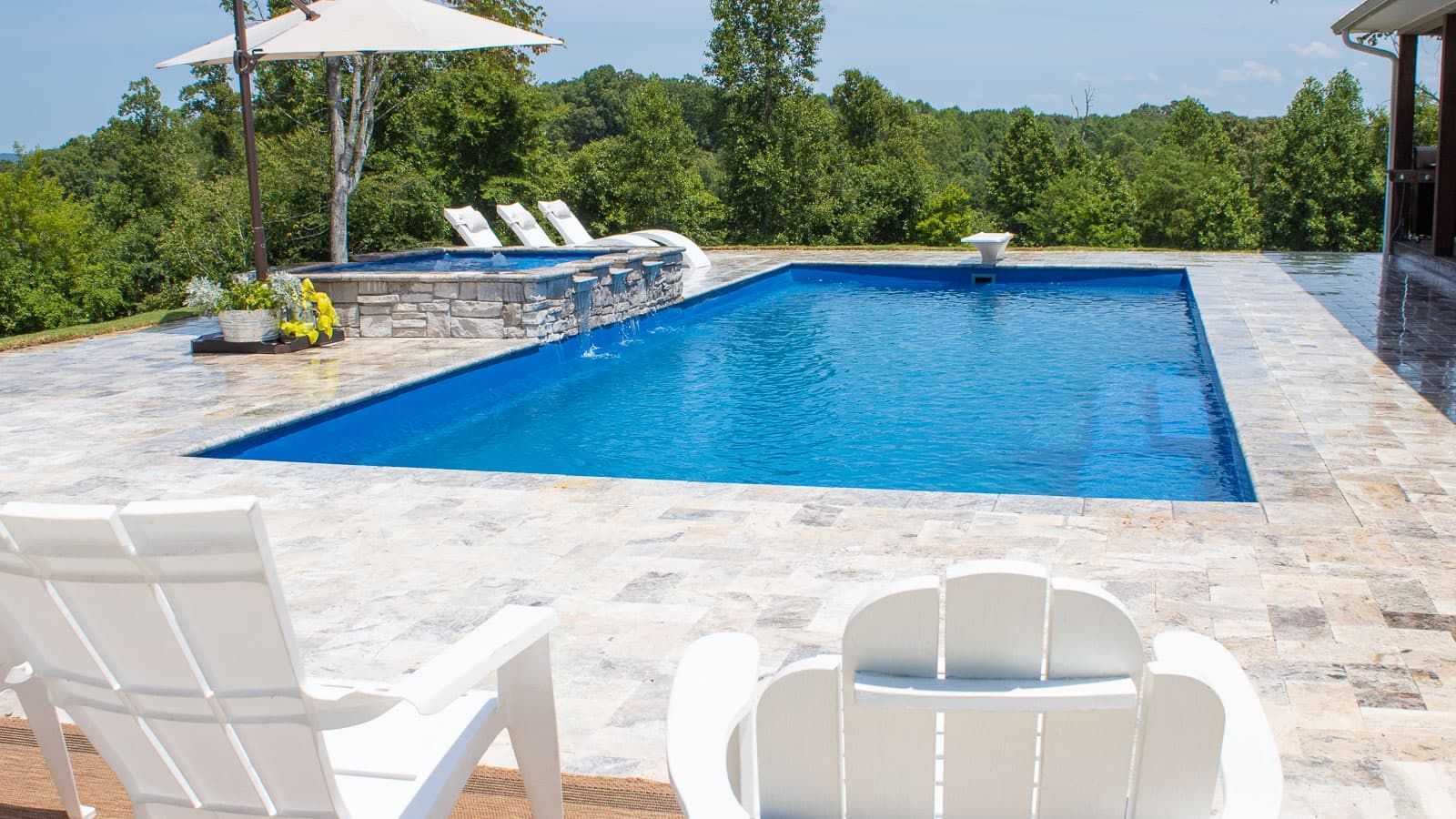 Backyard pool with blue water, white patio chairs, umbrella, and lounge chairs by greenery