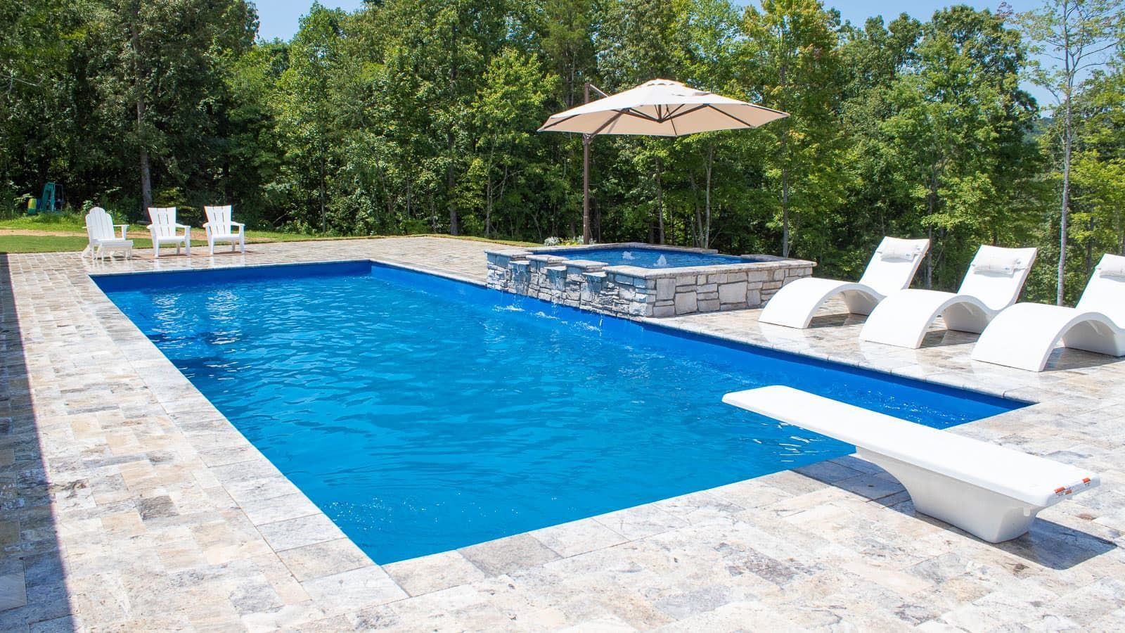 Rectangular blue swimming pool with stone deck, white lounge chairs, and umbrella beside trees