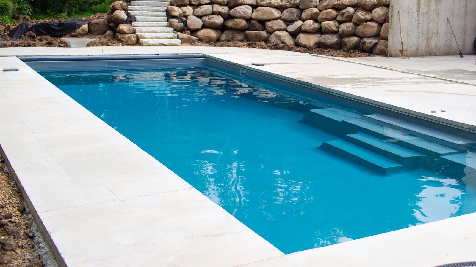 Rectangular blue swimming pool with white deck and stone wall backdrop