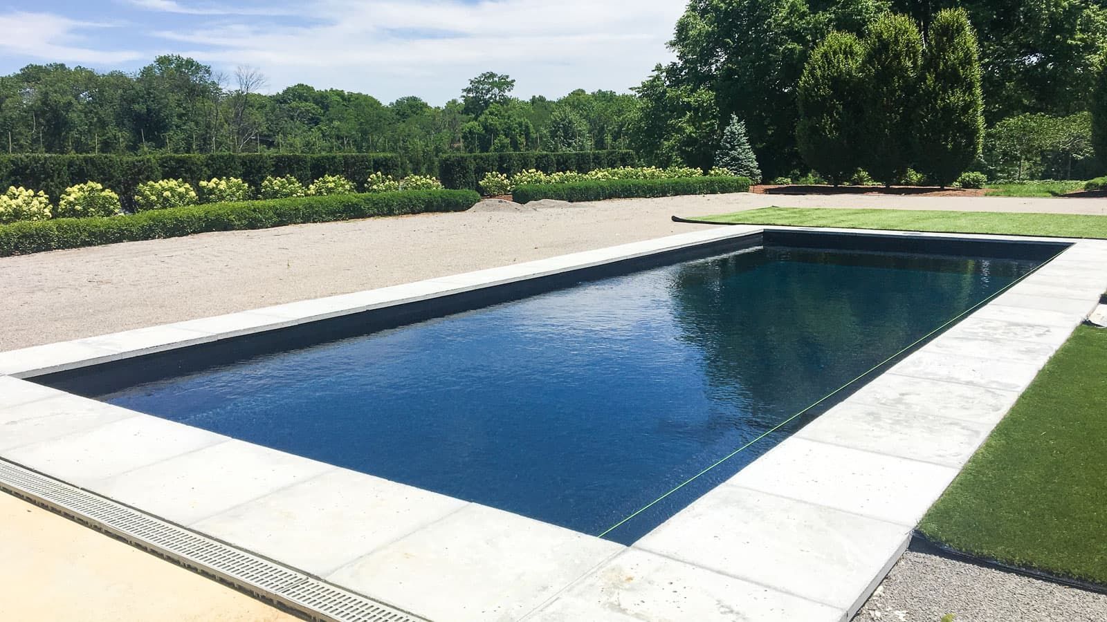 Rectangular outdoor swimming pool with white stone edging beside a green lawn and trees under a clear sky