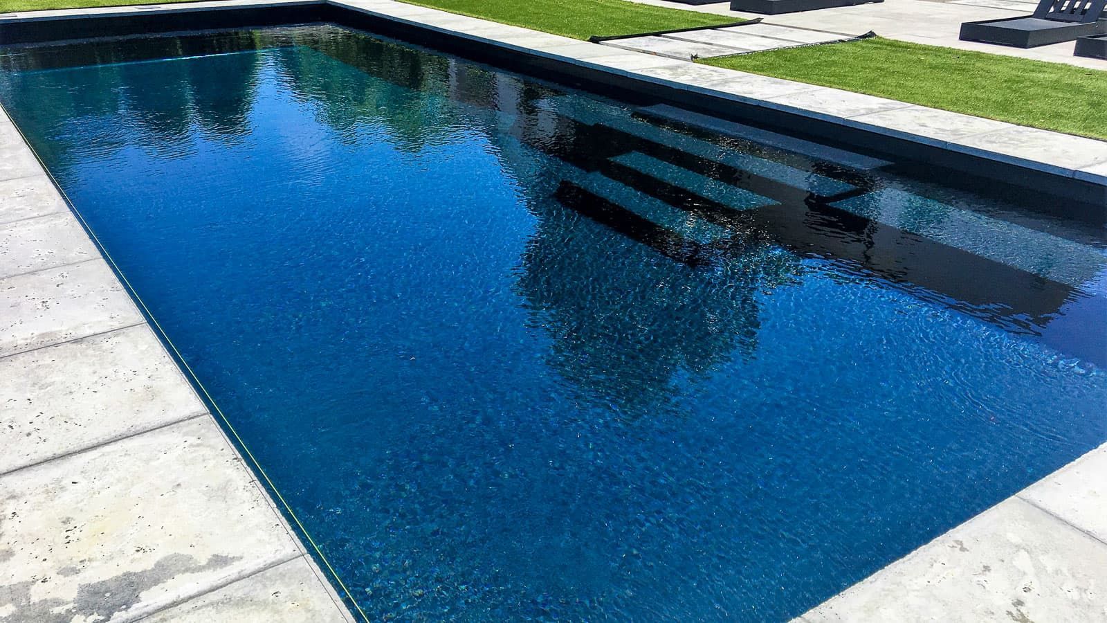 Rectangular blue swimming pool with stone deck and reflections of nearby trees and sky