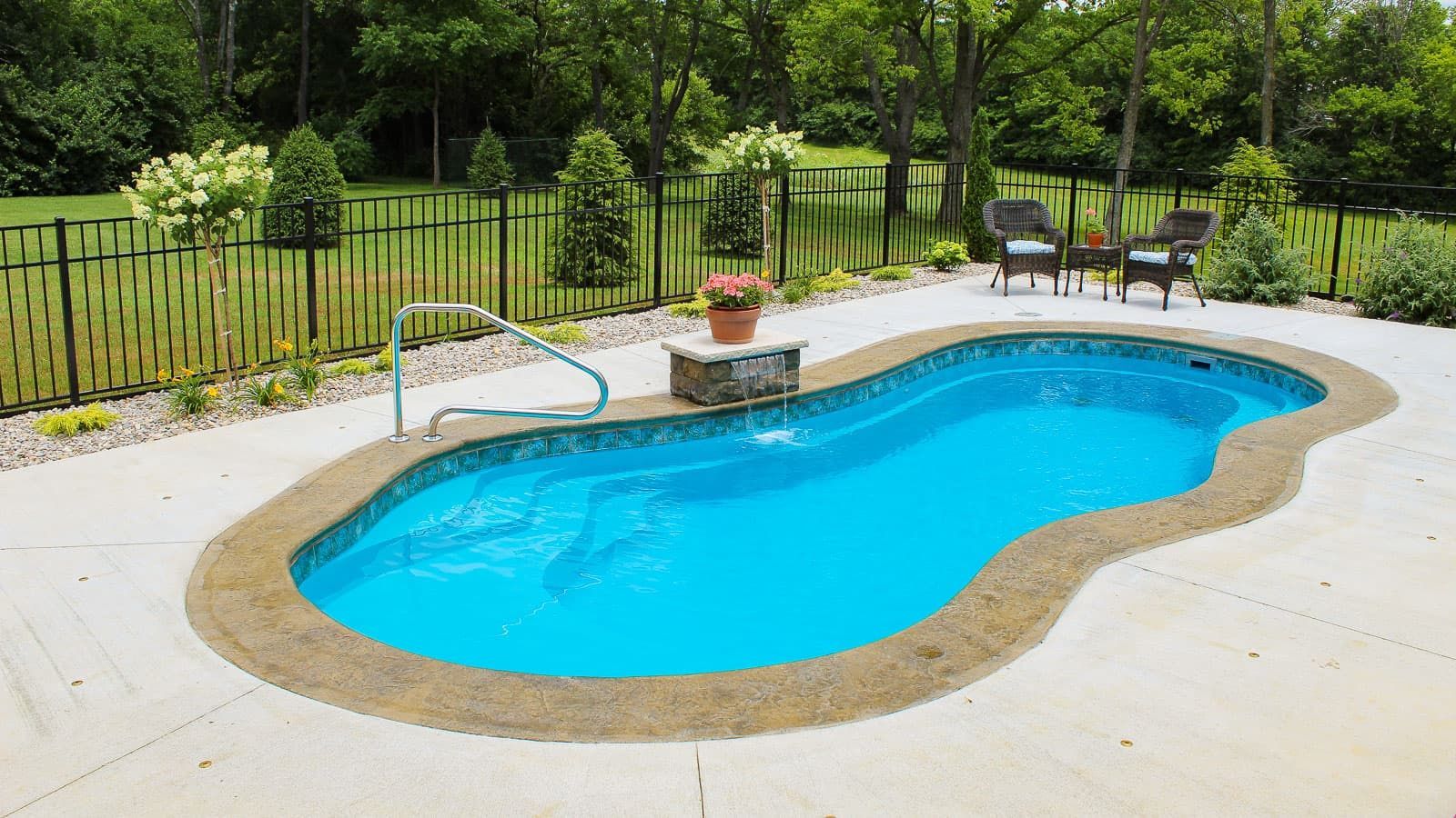 Backyard swimming pool with blue water, tan deck, fence, and patio chairs surrounded by greenery
