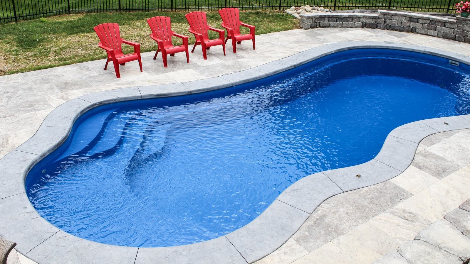 Backyard swimming pool with blue water, stone decking, and four red chairs on the patio