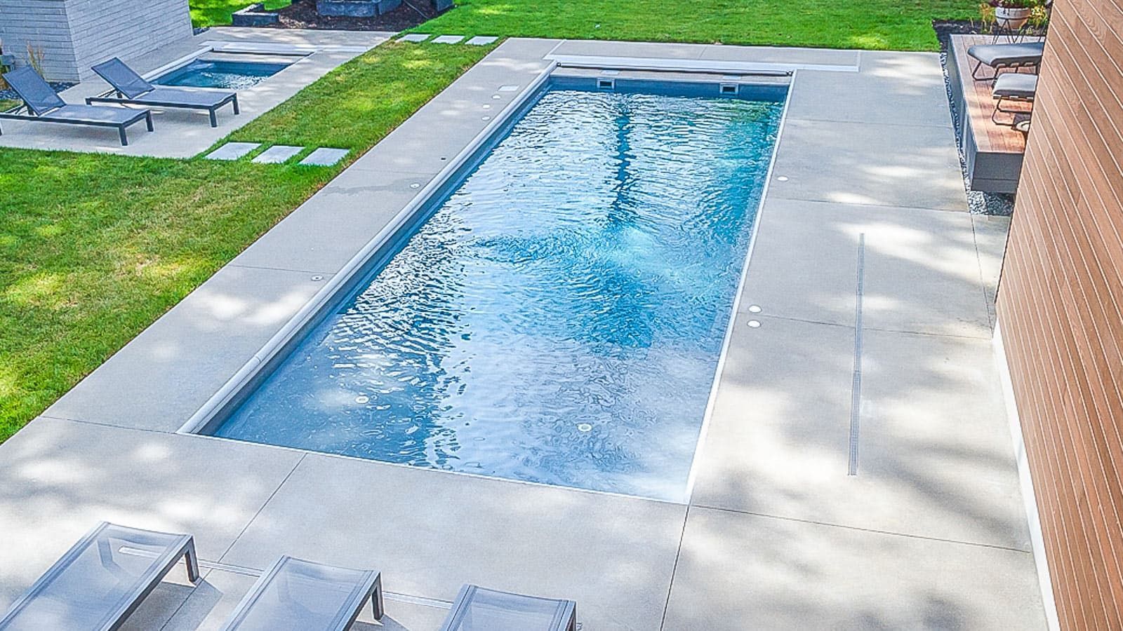 Rectangular backyard swimming pool with blue water, surrounded by a light patio and green lawn.
