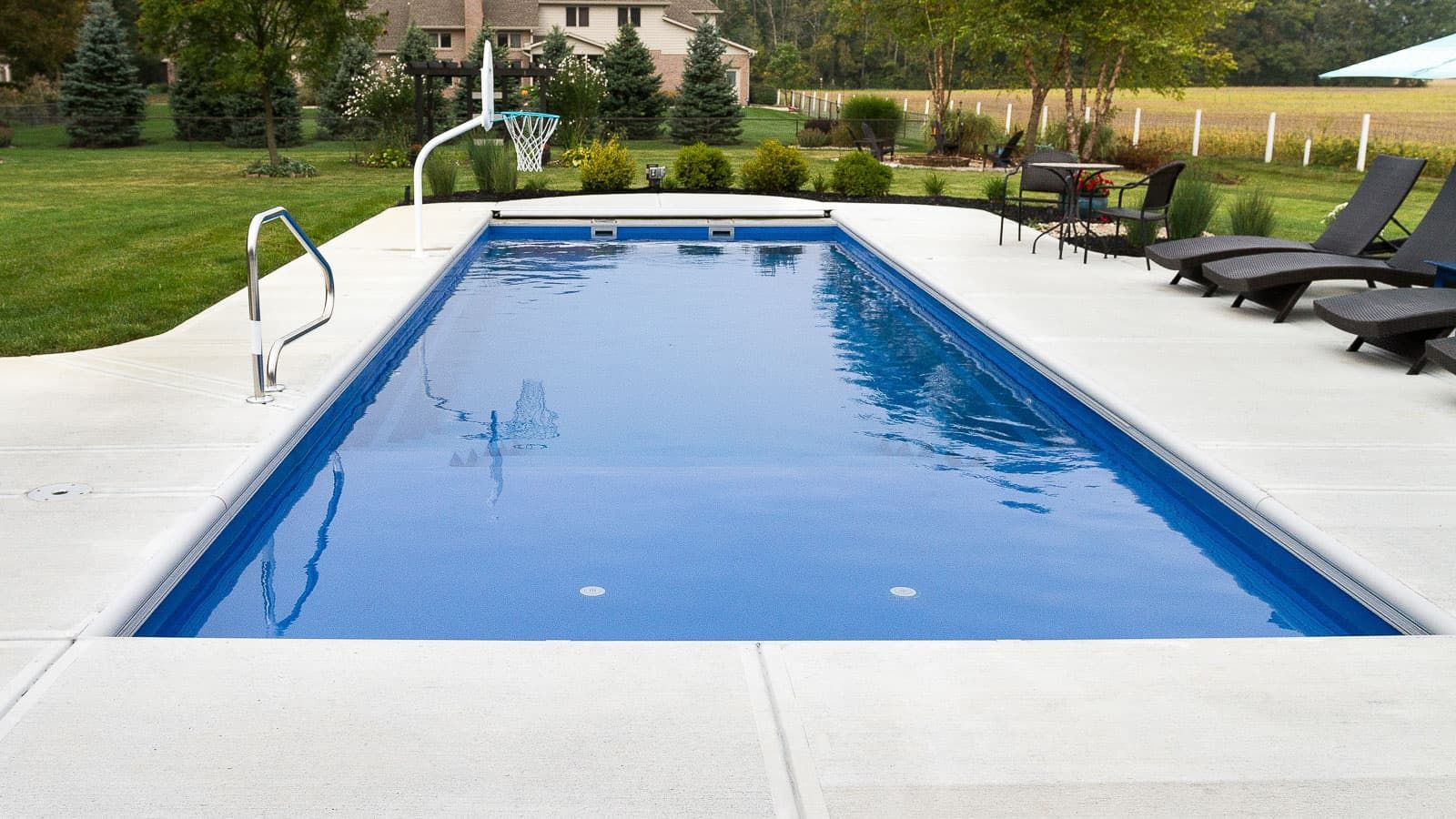 Rectangular blue swimming pool with white deck, handrails, and lounge chairs in a landscaped backyard