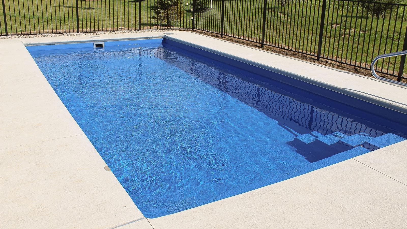 Rectangular backyard pool with blue water, white deck, and black fence behind it