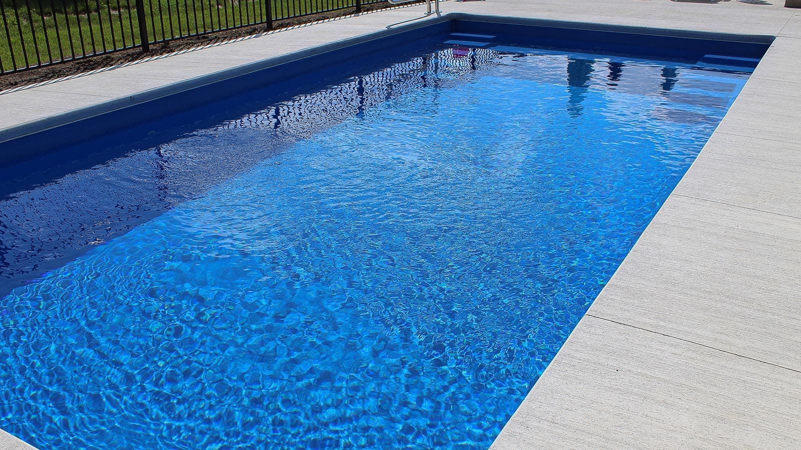 Rectangular blue swimming pool with white deck and black fence along the side