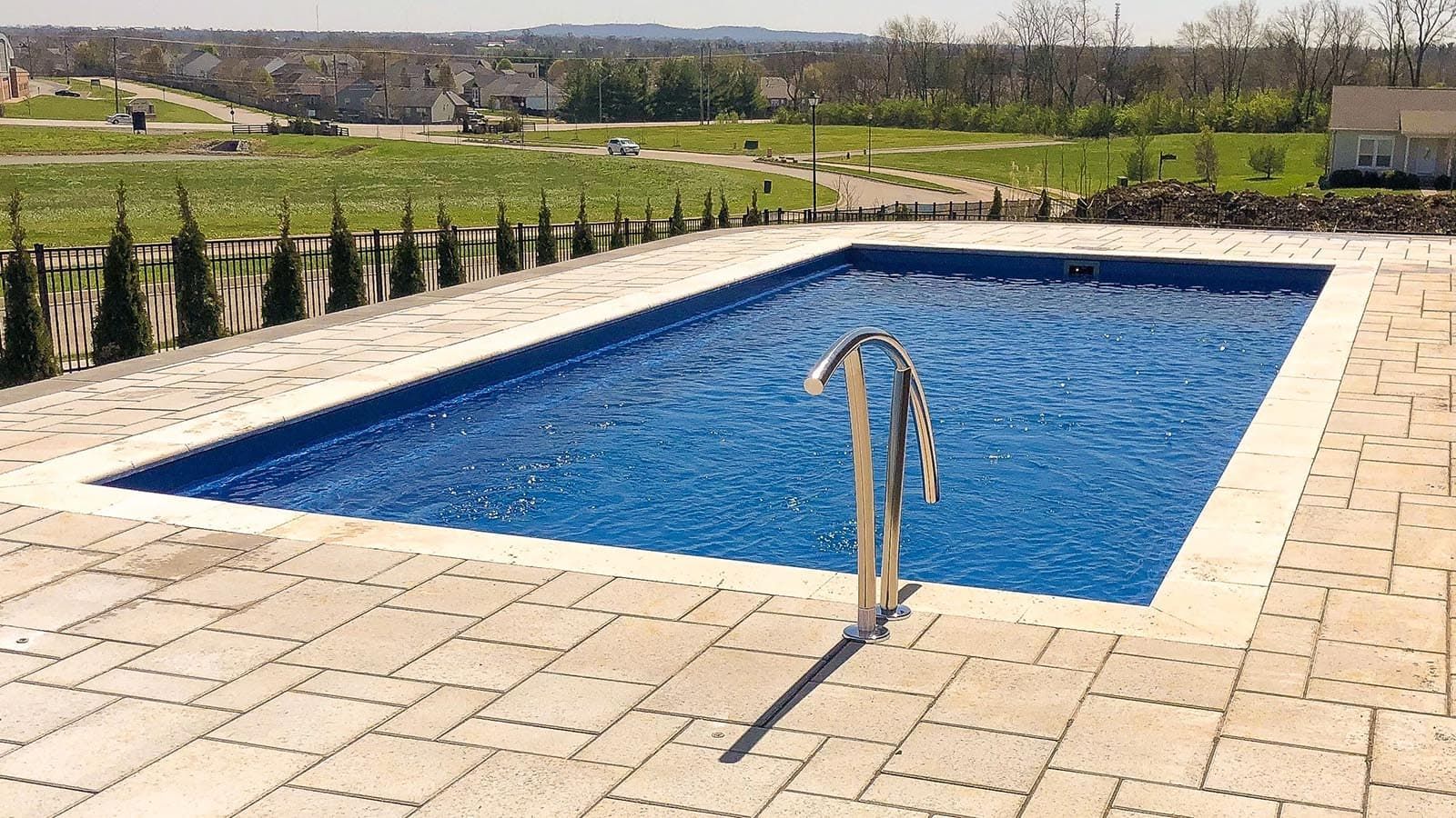 Rectangular blue swimming pool bordered by stone paving in a landscaped outdoor setting