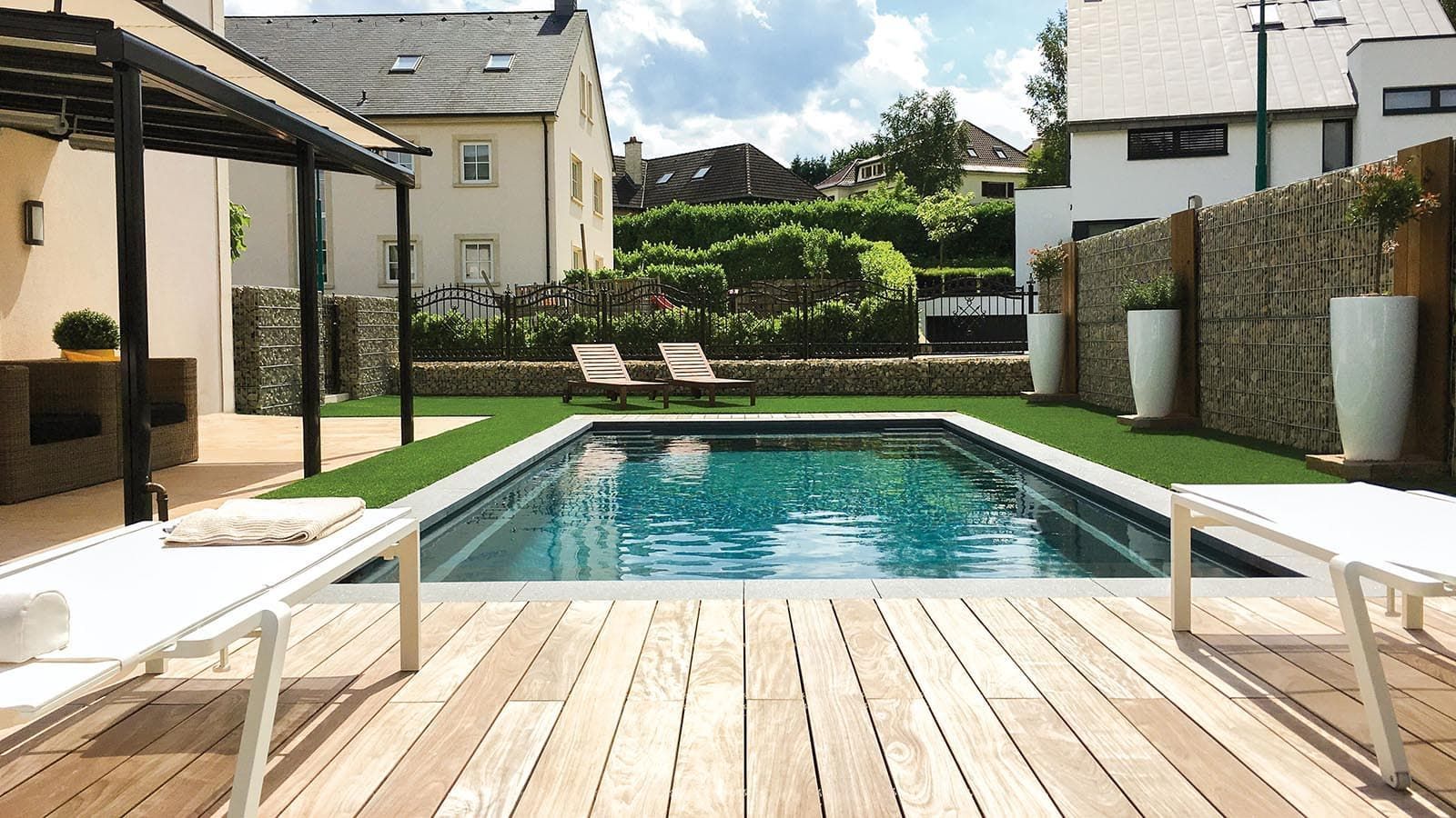 Backyard pool with wooden deck, lounge chairs, and modern houses on a sunny day