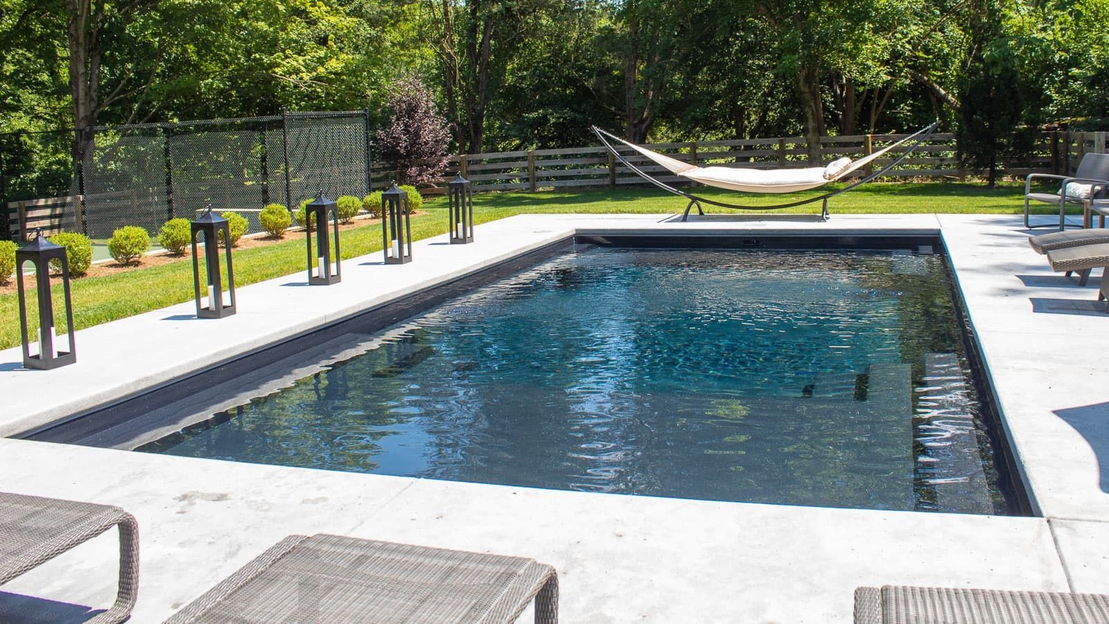 Backyard pool with a hammock, lounge chairs, and green trees beyond the fence