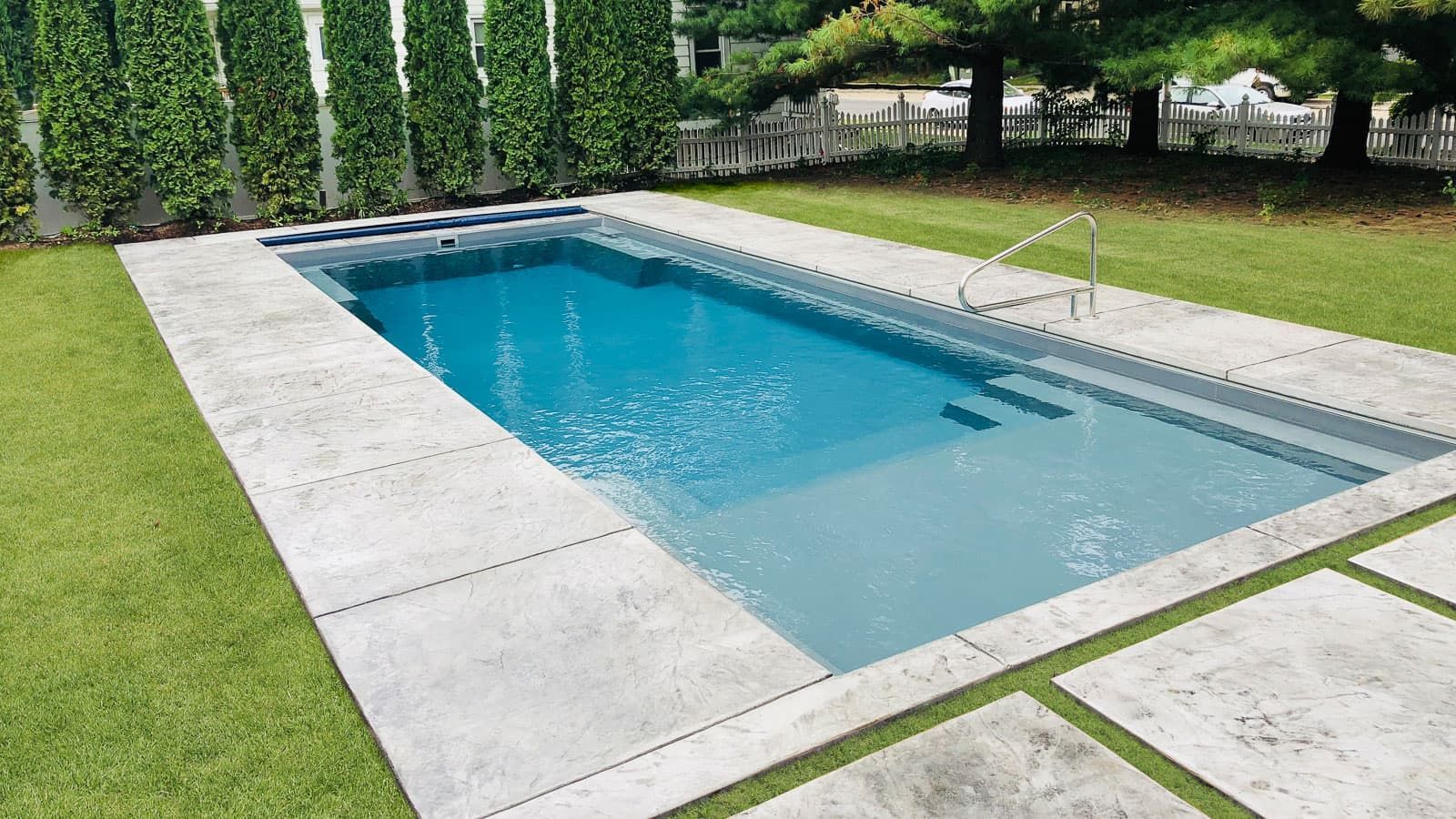 Rectangular backyard swimming pool with blue water, concrete deck, green lawn, and tall hedge fence