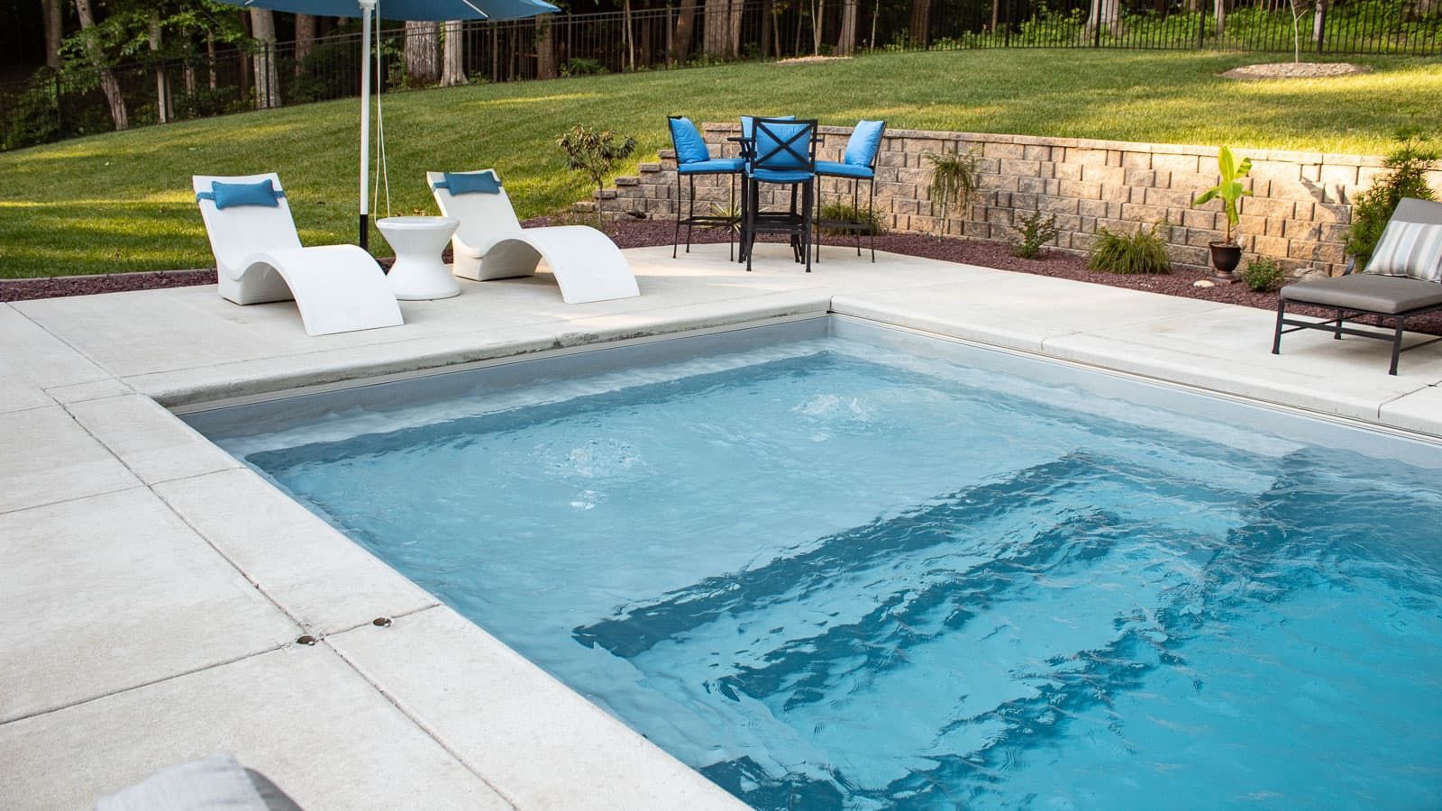 Backyard pool with blue water, white lounge chairs, and a patio table by a stone wall and trees