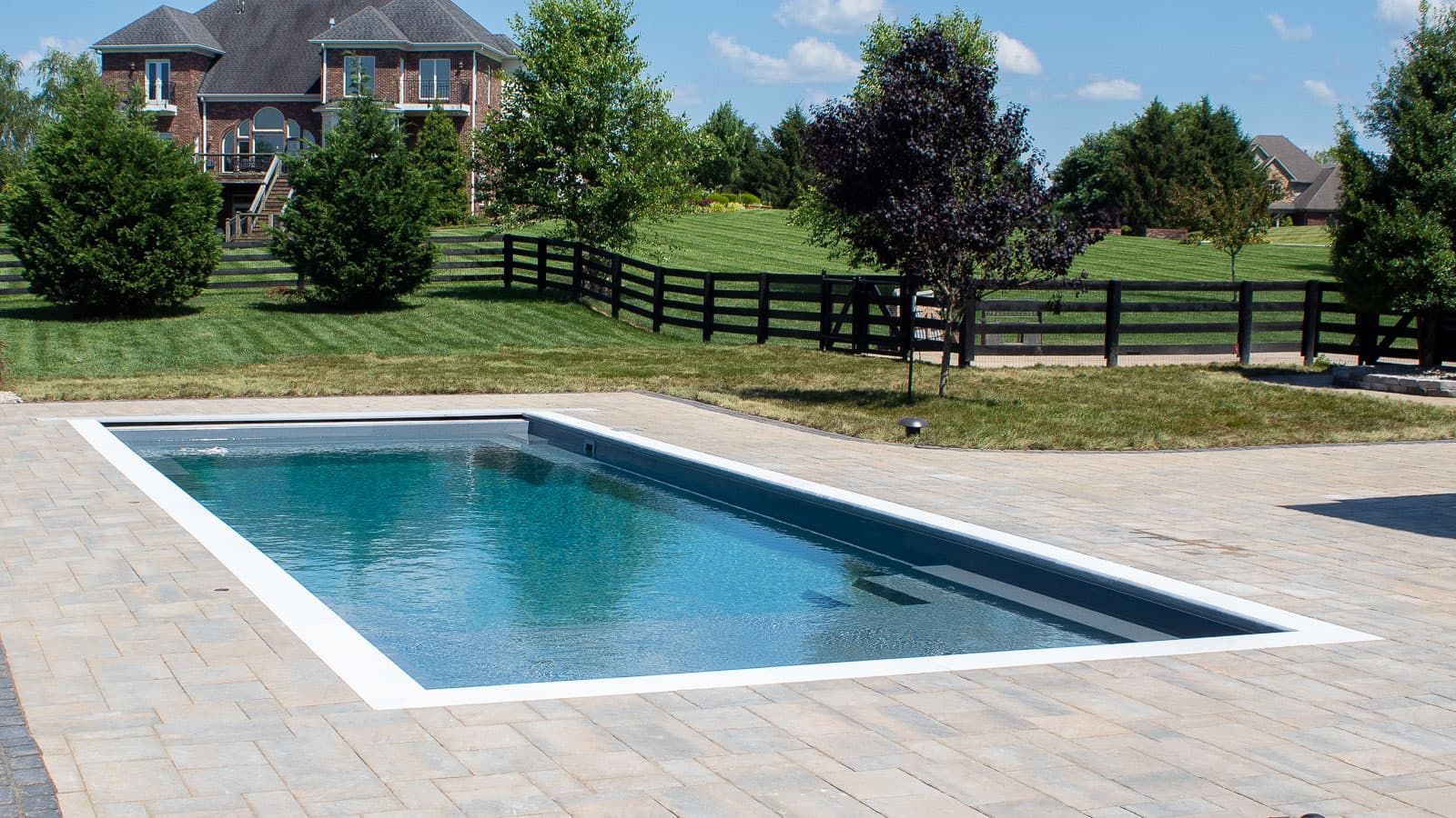 Rectangular backyard swimming pool with blue water beside a stone patio, fence, trees, and houses in the background