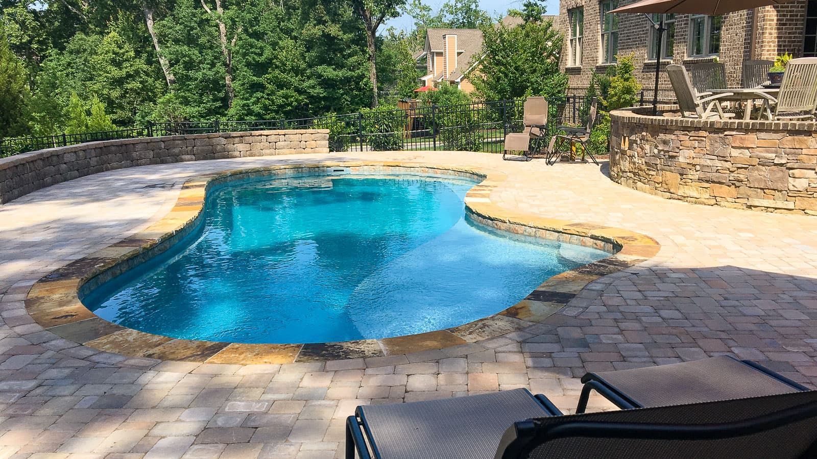 Curved backyard swimming pool with blue water, stone patio, and patio chairs in a sunny landscaped yard