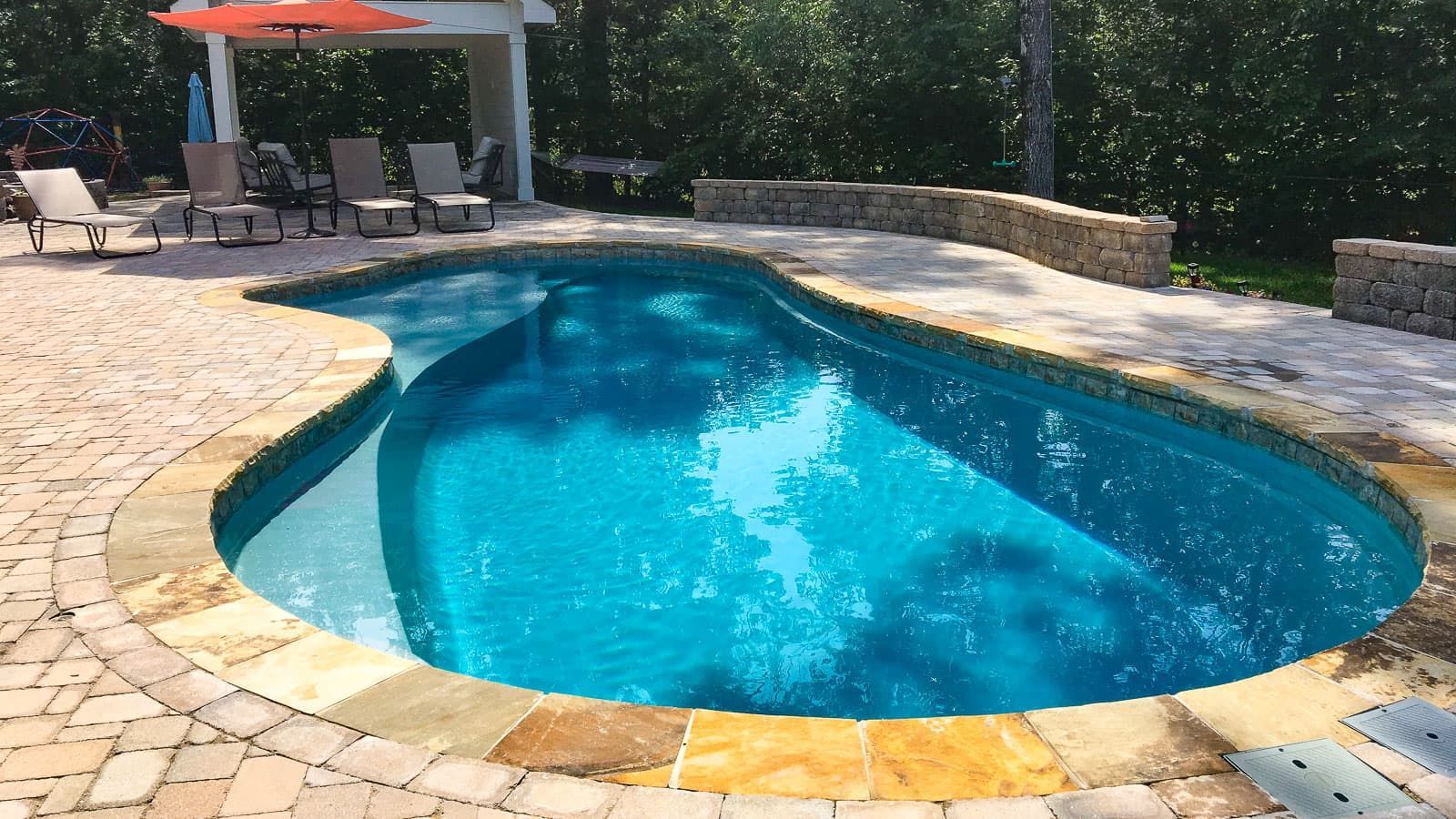 Curved backyard swimming pool with blue water, stone coping, and patio seating under an umbrella