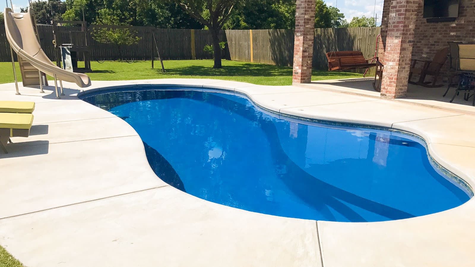 Curved backyard swimming pool with bright blue water beside a patio and brick house