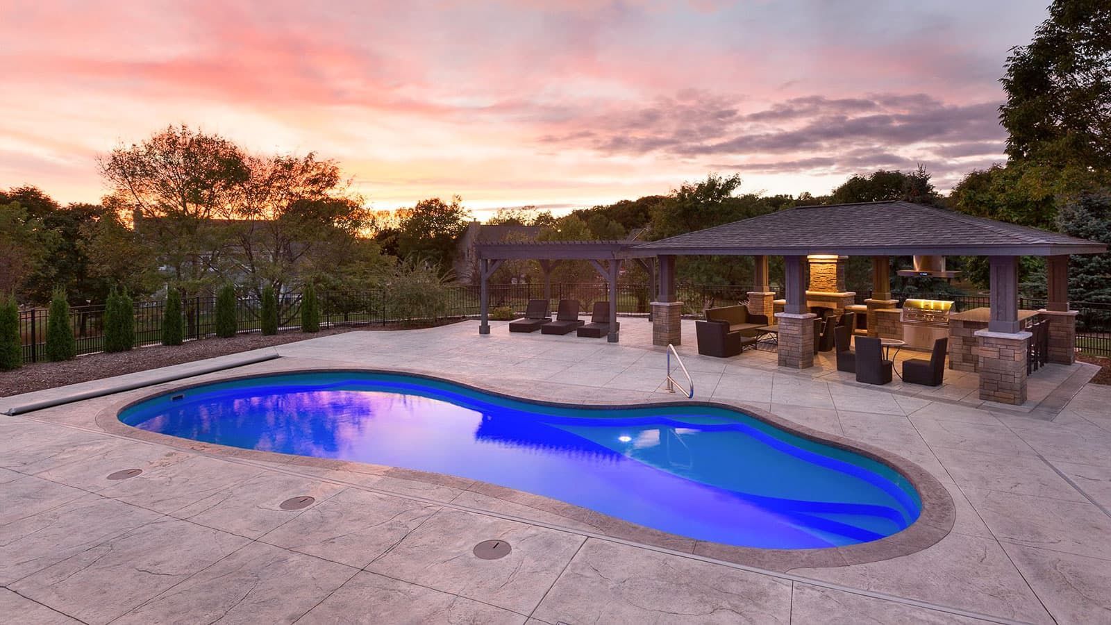 Backyard pool glowing blue at sunset beside a lit patio and outdoor dining area