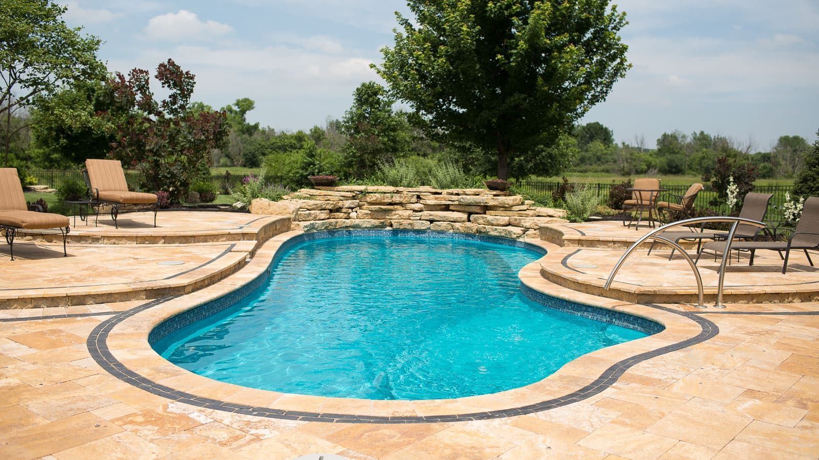 Backyard swimming pool with tan stone deck and lounge chairs, surrounded by trees and greenery