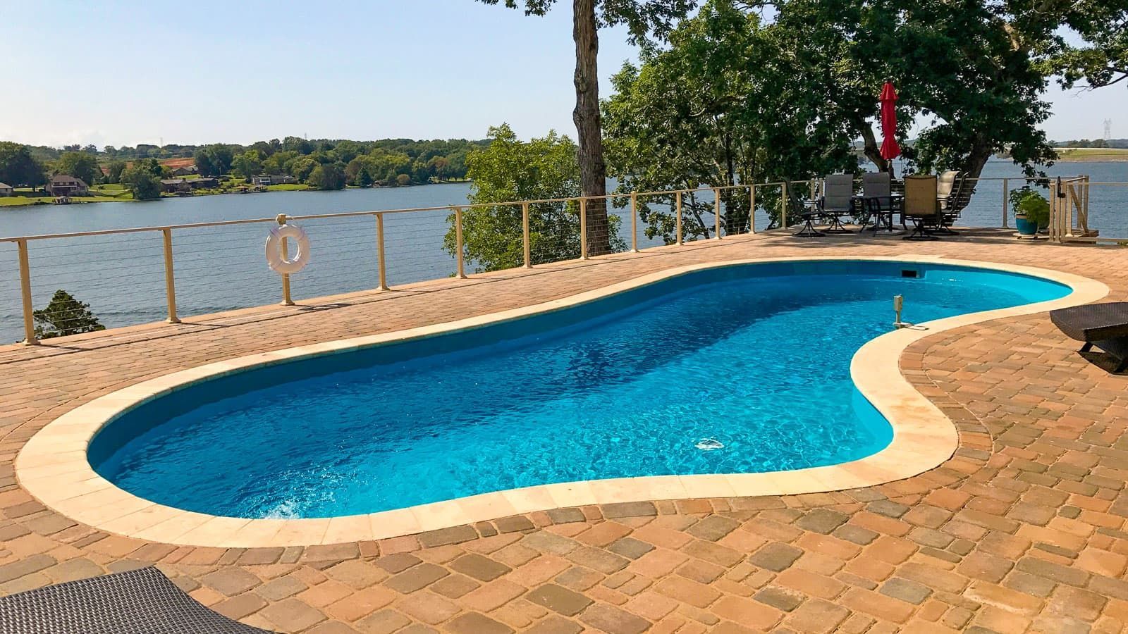 Curved blue swimming pool beside a brick patio, overlooking a lake on a sunny day
