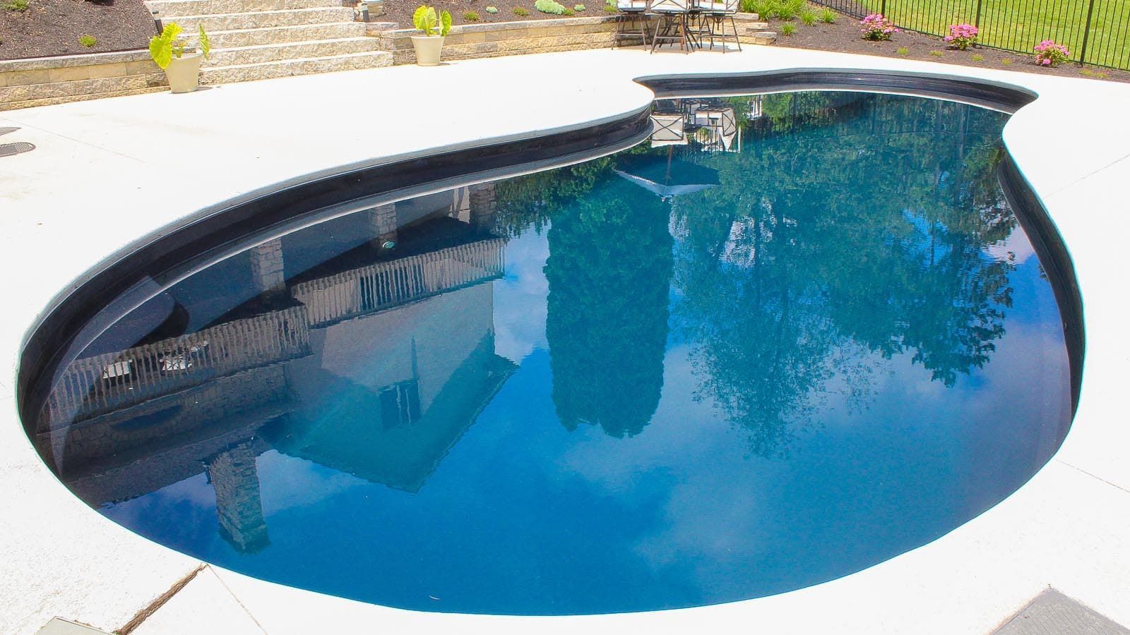 Curved blue swimming pool with white stone deck and a wall reflection on the water