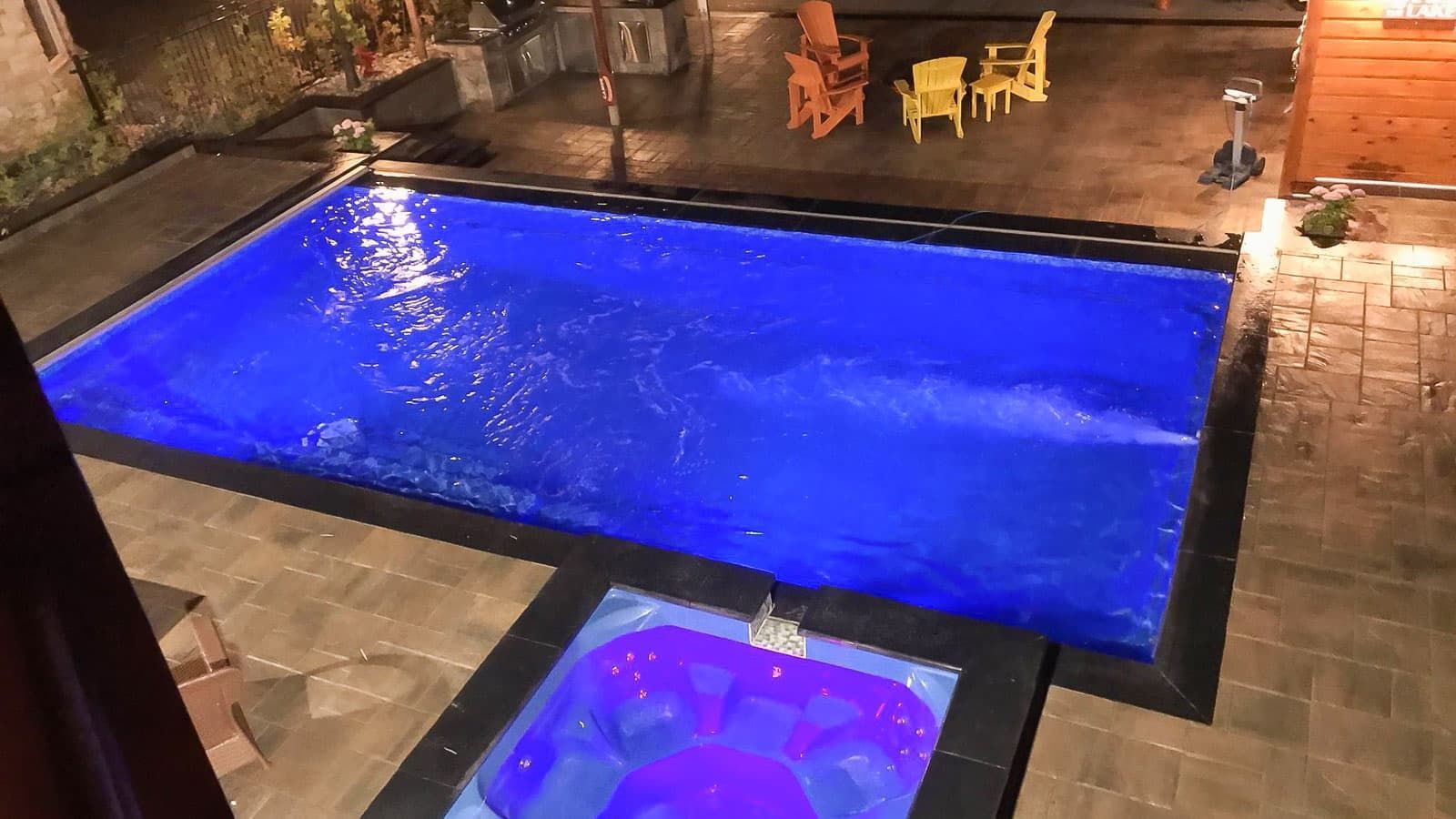 Nighttime backyard with a lit rectangular pool and small hot tub, both glowing blue and purple