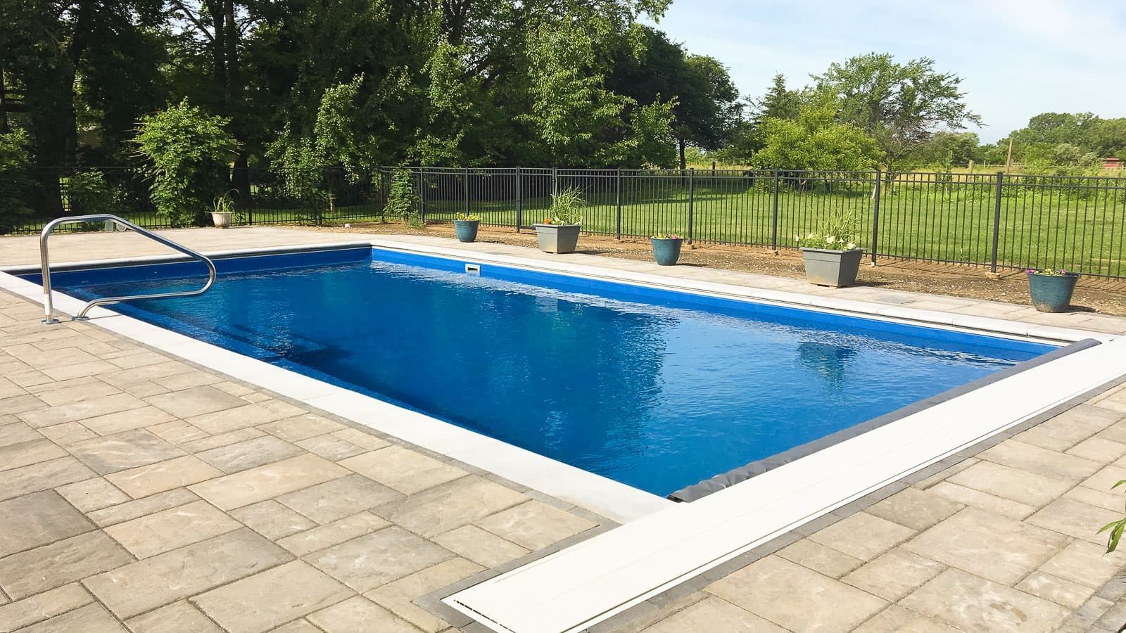 Backyard swimming pool with blue water, white coping, and a stone patio beside a grassy fence line